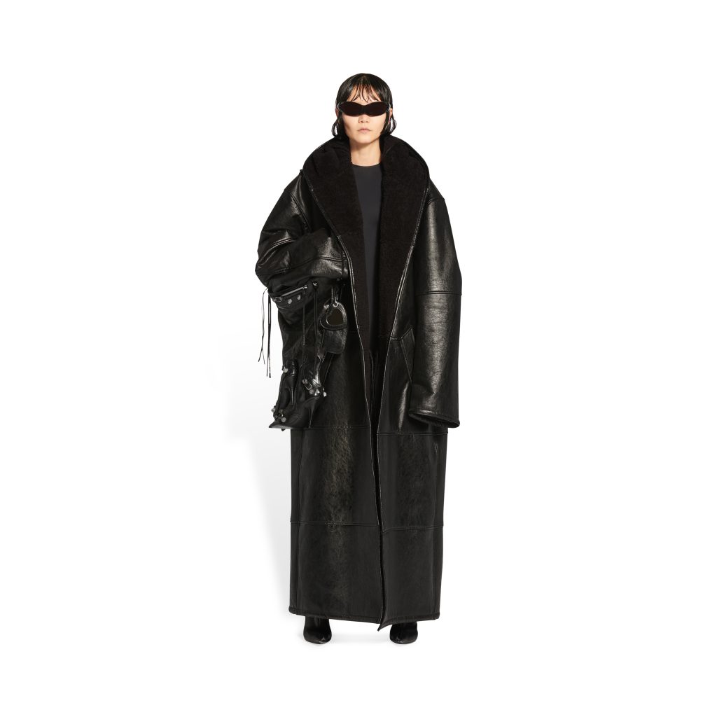 Model wearing an oversized black leather coat with a shearling collar and a studded bag, Balenciaga