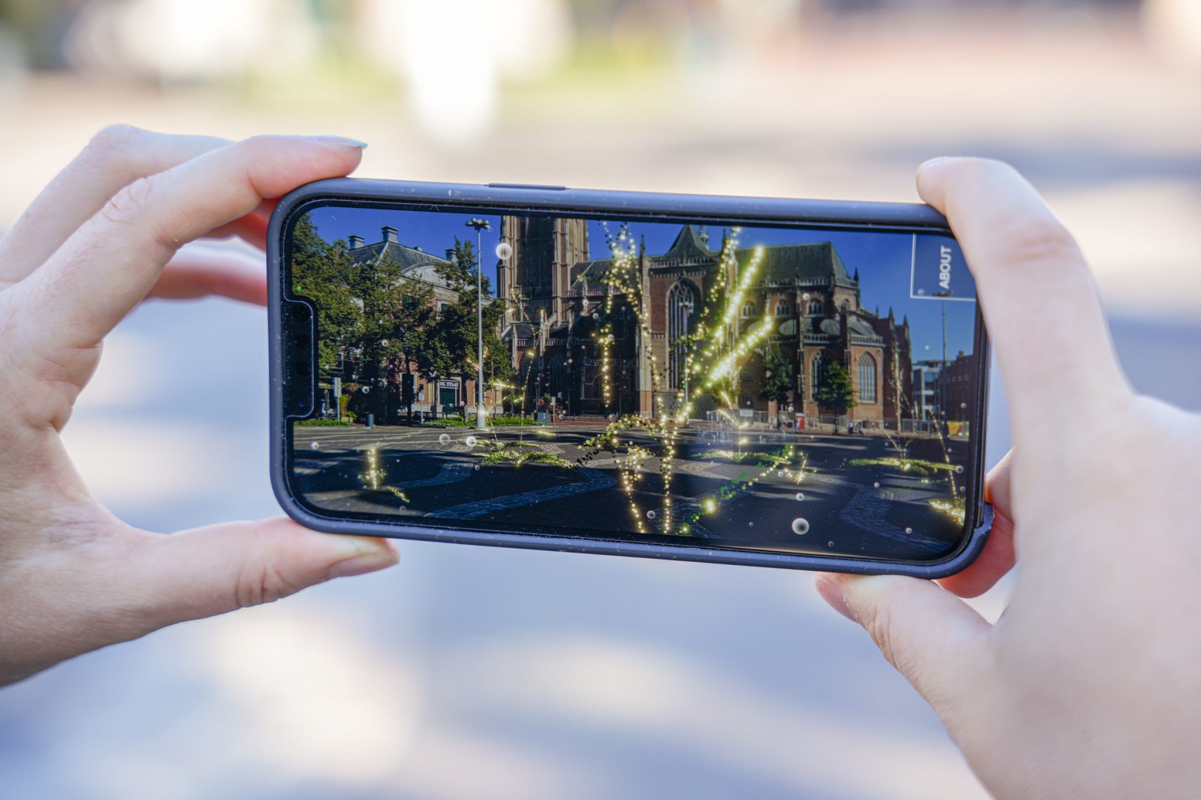 Smartphone screen shows the “Arabidopsis Symphony” AR installation with glowing digital plants over a church square