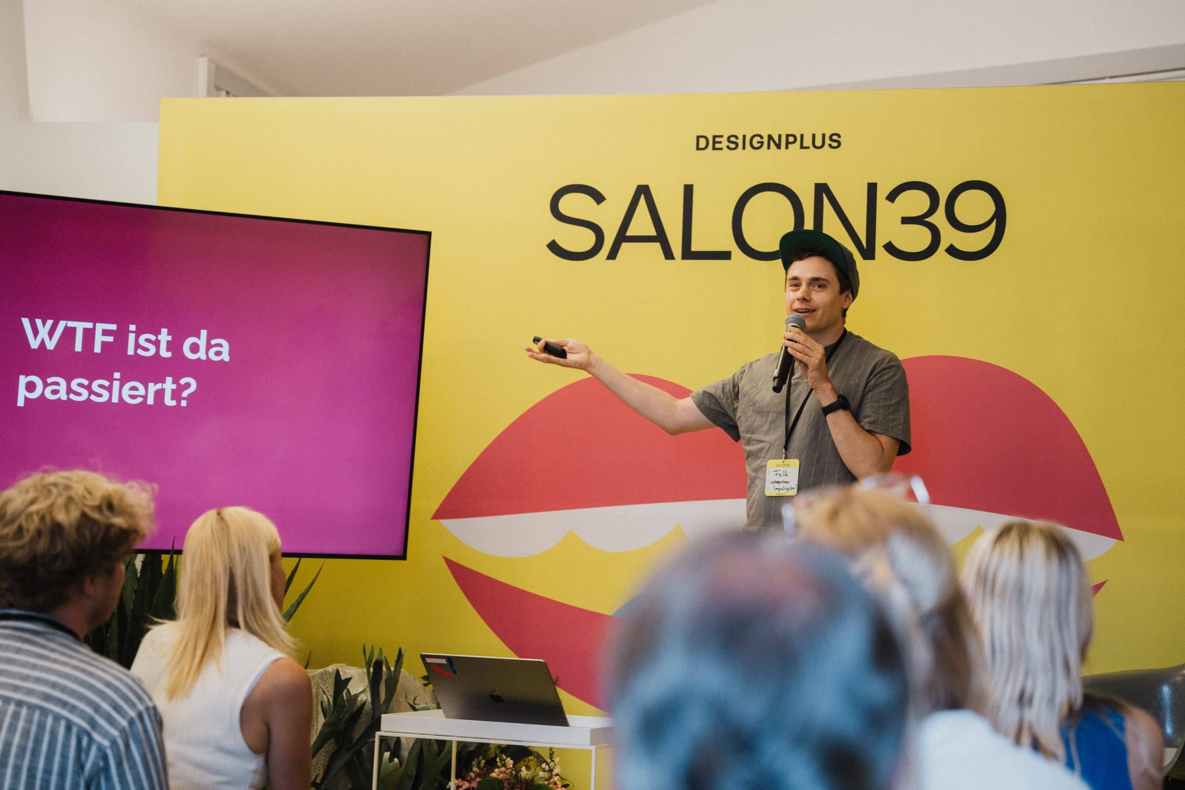Speaker vor gelber SALON39-Bühnenwand mit Lippen-Motiv, daneben Slide „WTF ist da passiert?“