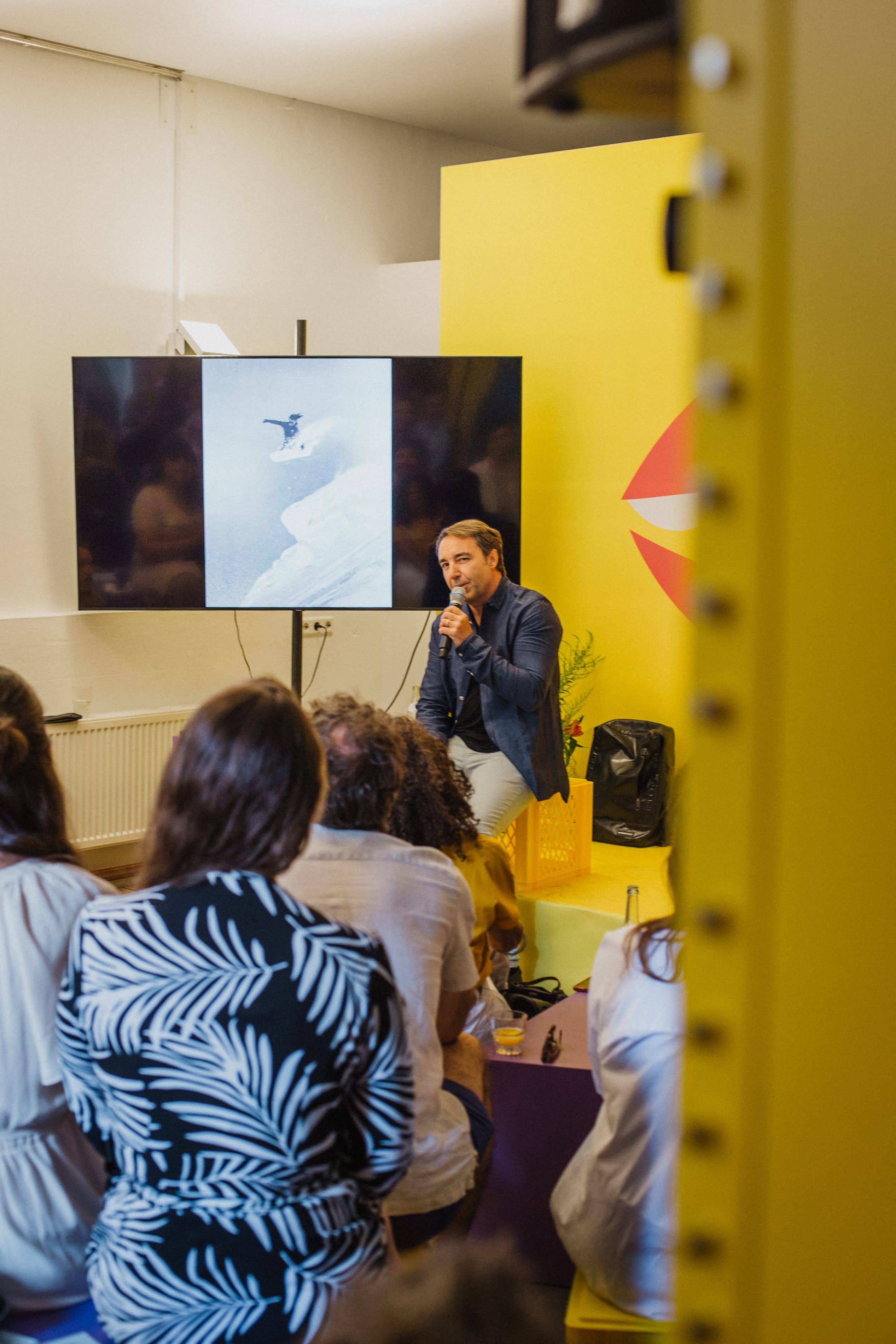 Speaker mit Mikrofon vor Leinwand mit Surf-Foto, gelbe Bühnenwand im Hintergrund
