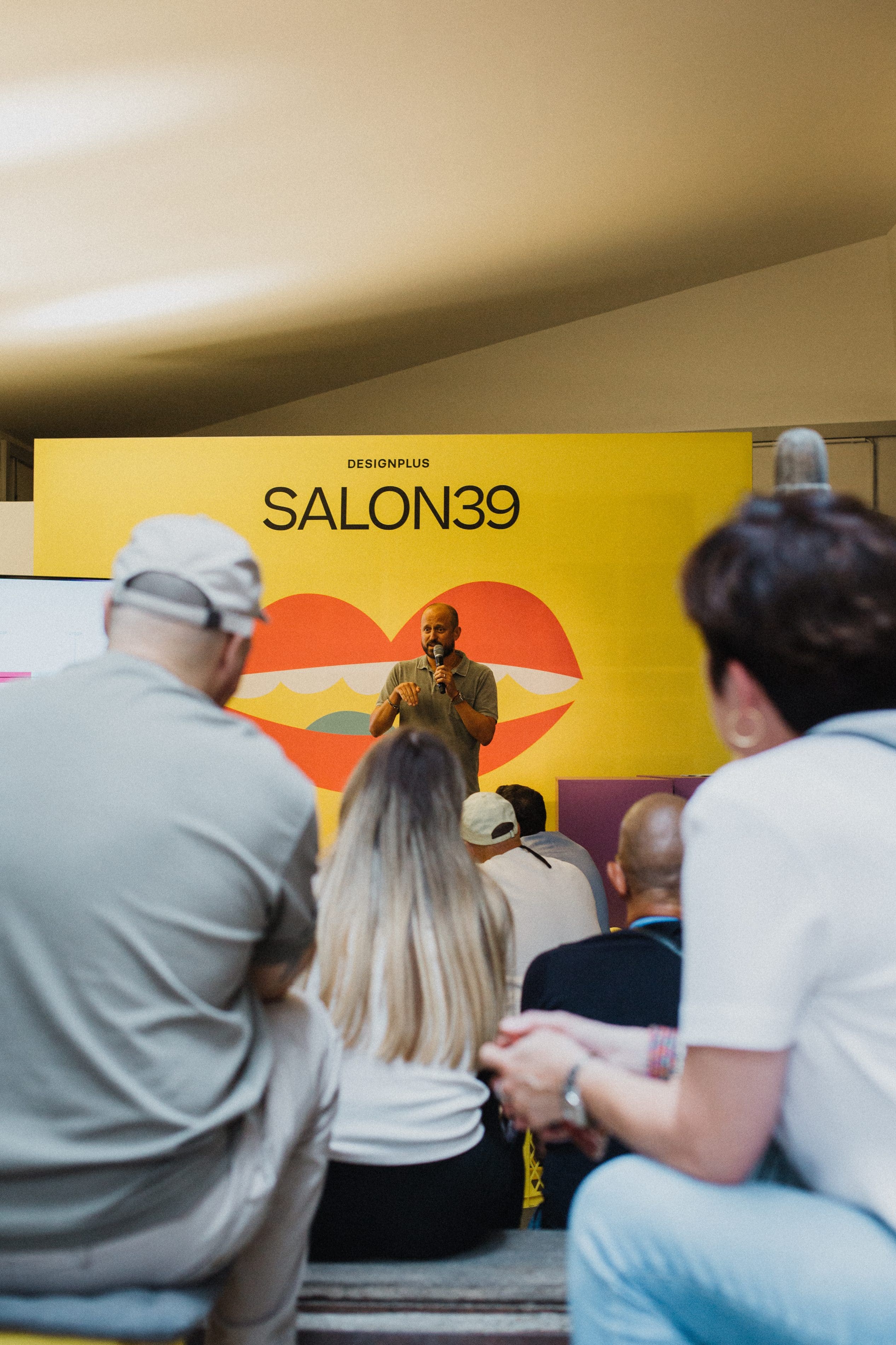 Speaker mit Mikrofon vor gelbem SALON39-Banner mit Lippen-Illustration, Publikum im Vordergrund