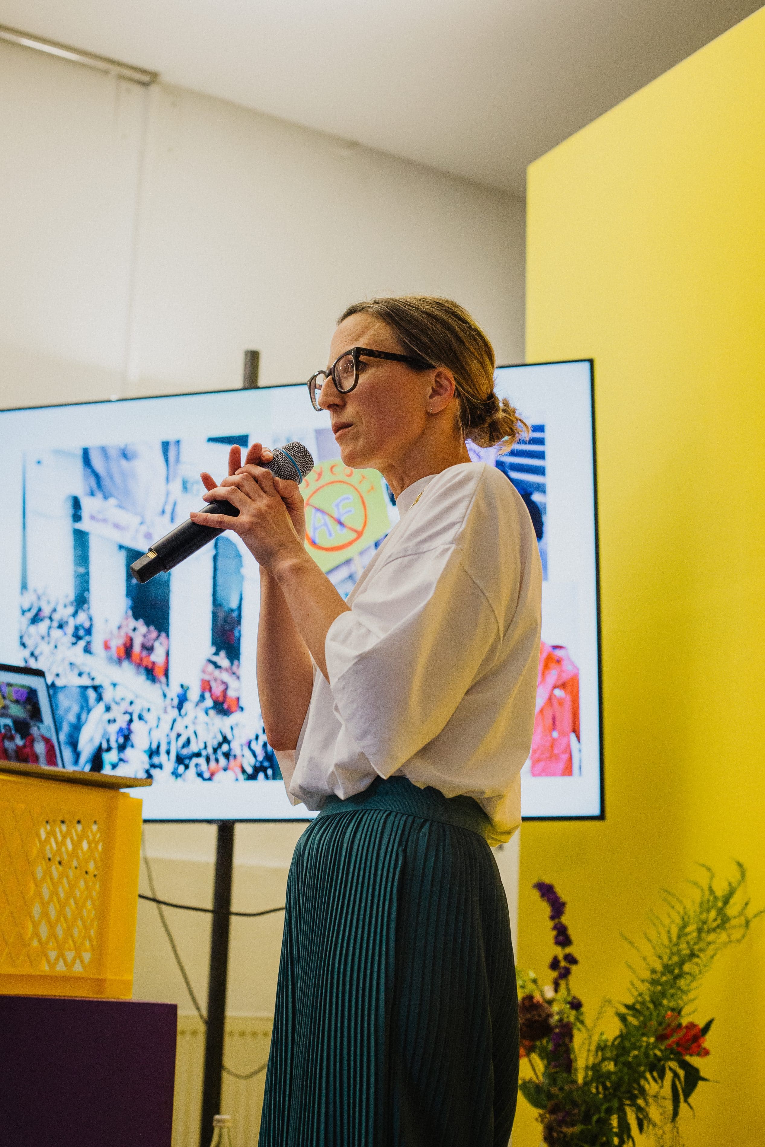Speakerin mit Mikrofon vor Bildschirm mit Menschenmenge-Foto, gelbe Wand im Hintergrund