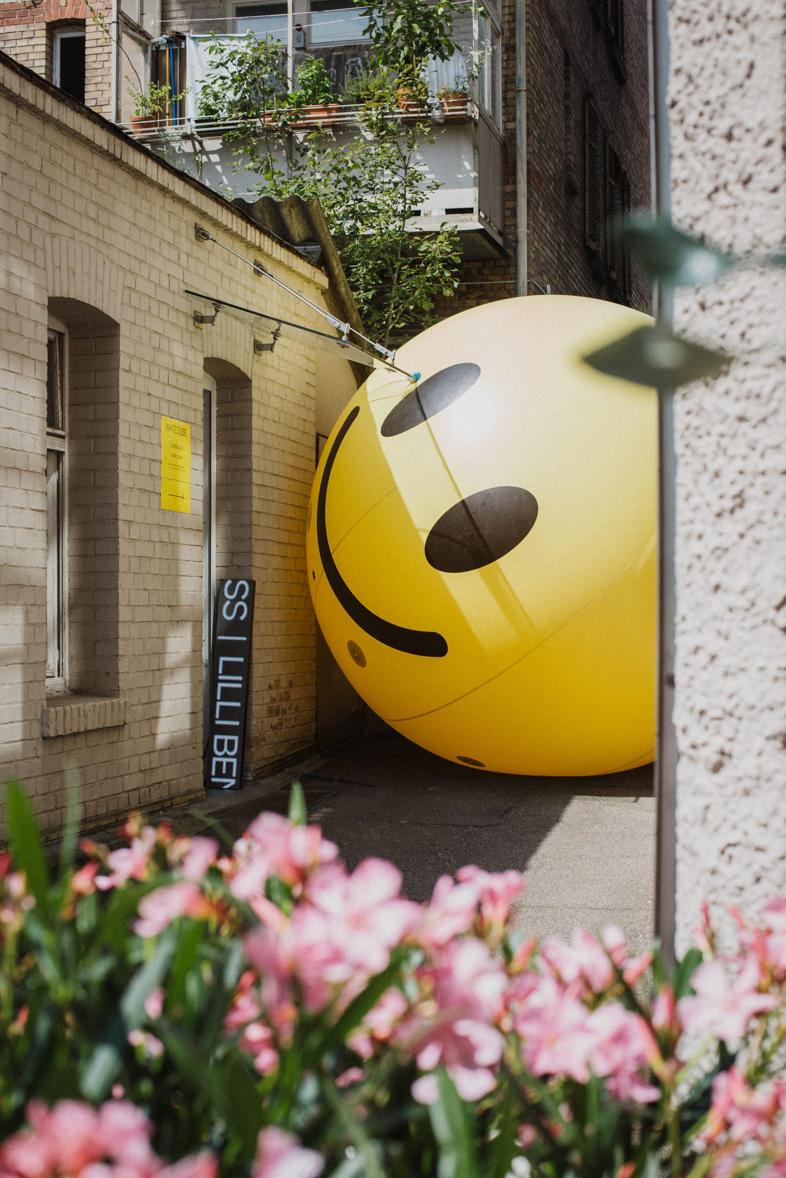 Riesiger gelber Smiley-Ballon zwischen Hinterhofgebäuden, im Vordergrund rosa Blüten