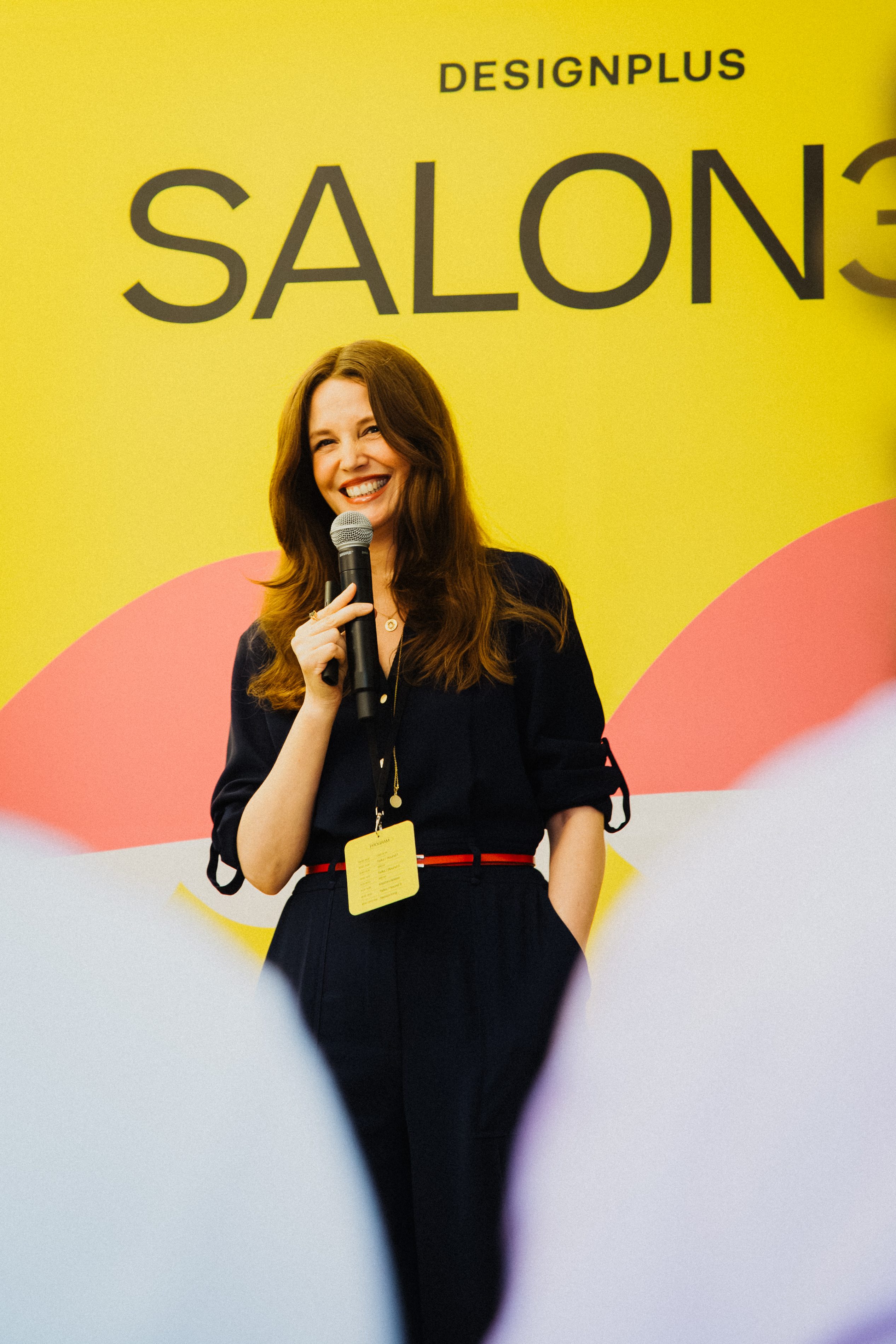 A speaker holds a microphone in front of the yellow SALON39 stage backdrop by Designplus.