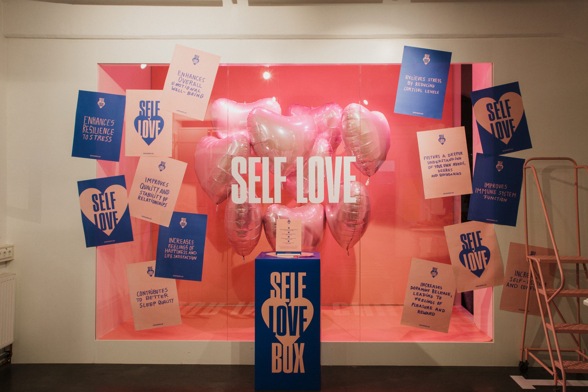 A “Self Love” window installation with pink heart-shaped balloons and information cards.