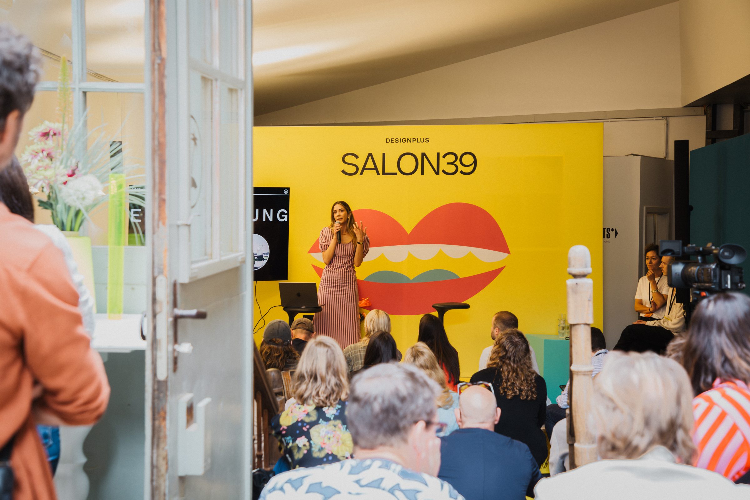 A speaker on stage in front of the lips-motif SALON39 backdrop, with the audience seated in front.
