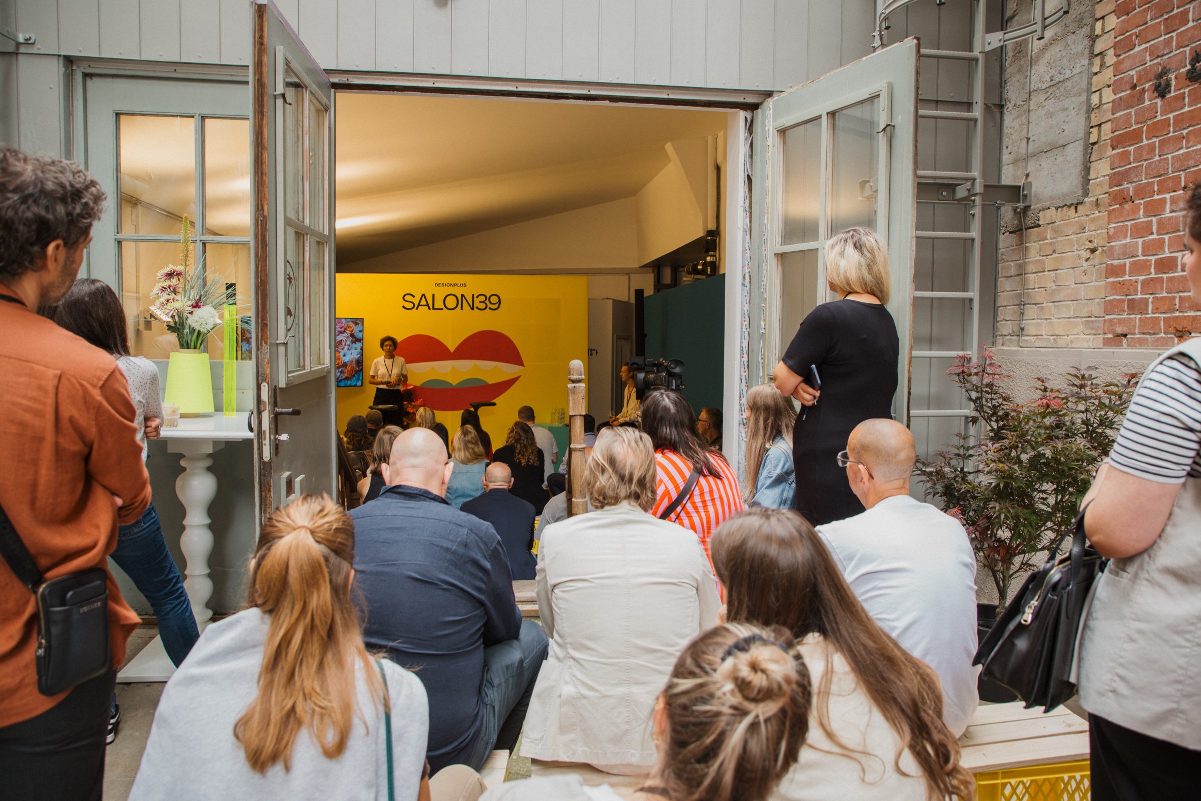 The audience sits outside the doorway watching a talk inside the yellow SALON39 room.