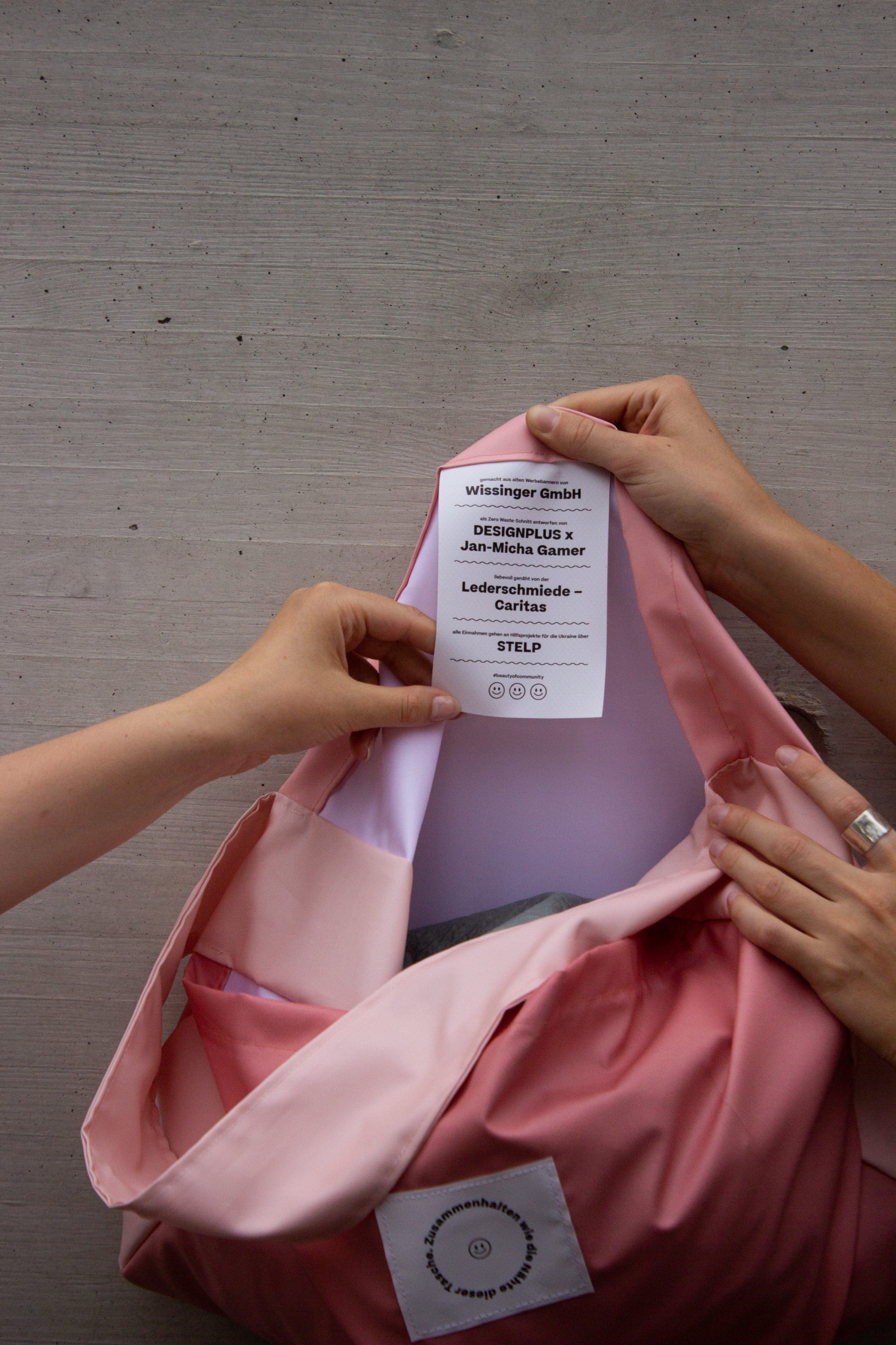 Hands open a pink Charity Bag revealing the label crediting Designplus x Jan-Micha Gamer and Stelp.