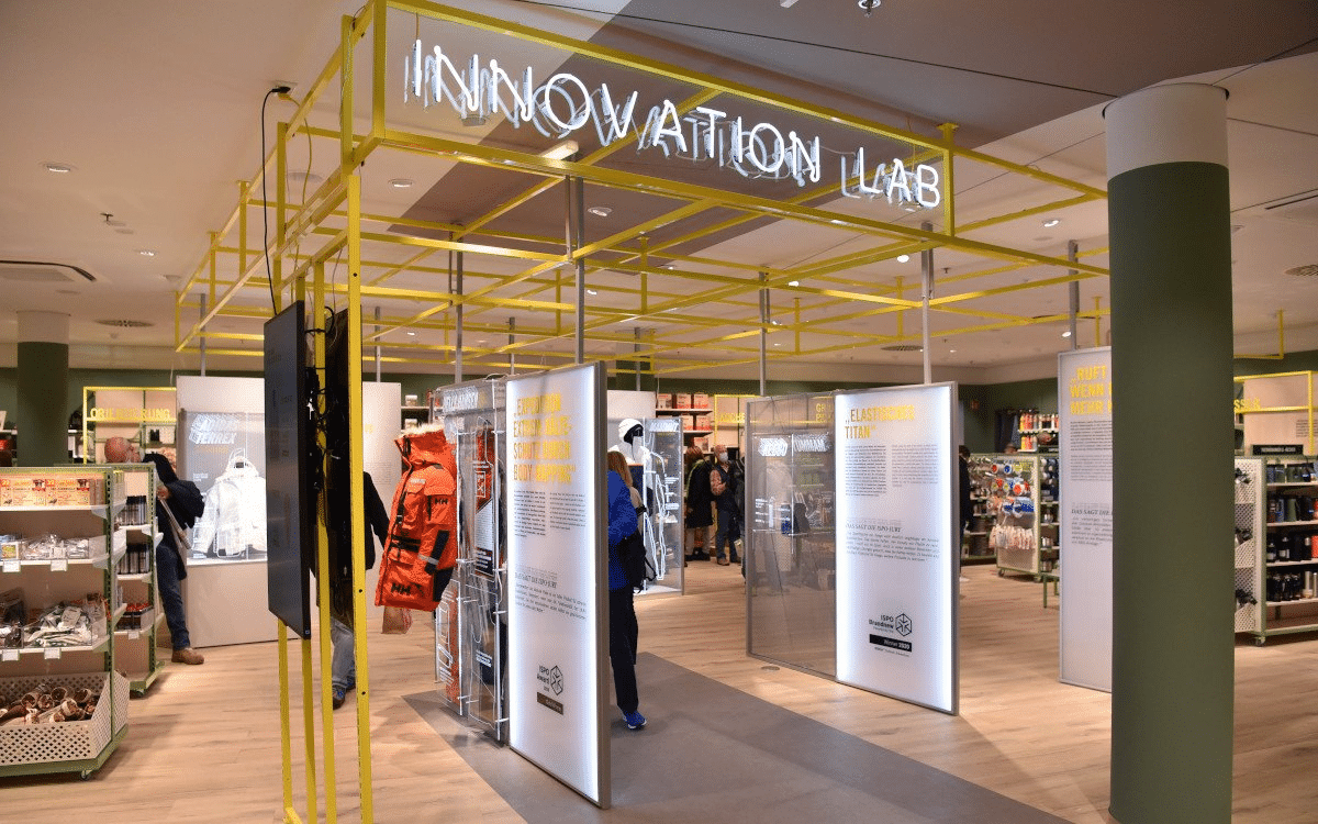 „Innovation Lab“-Bereich mit gelbem Rohrgestell an der Decke und beleuchteten Infotafeln im Store.