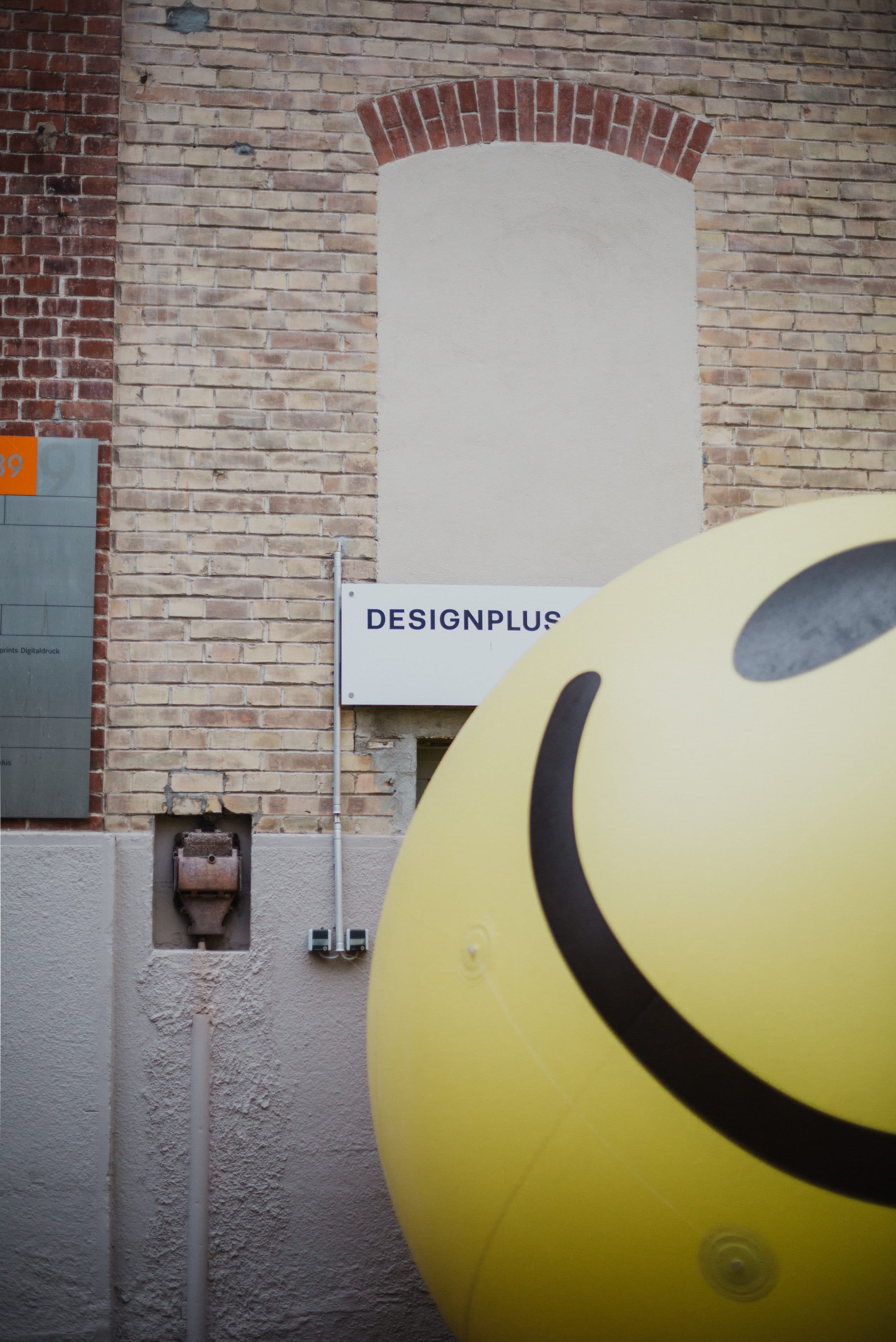 Gelber Smiley-Ballon vor der Backsteinfassade mit „Designplus“-Schild