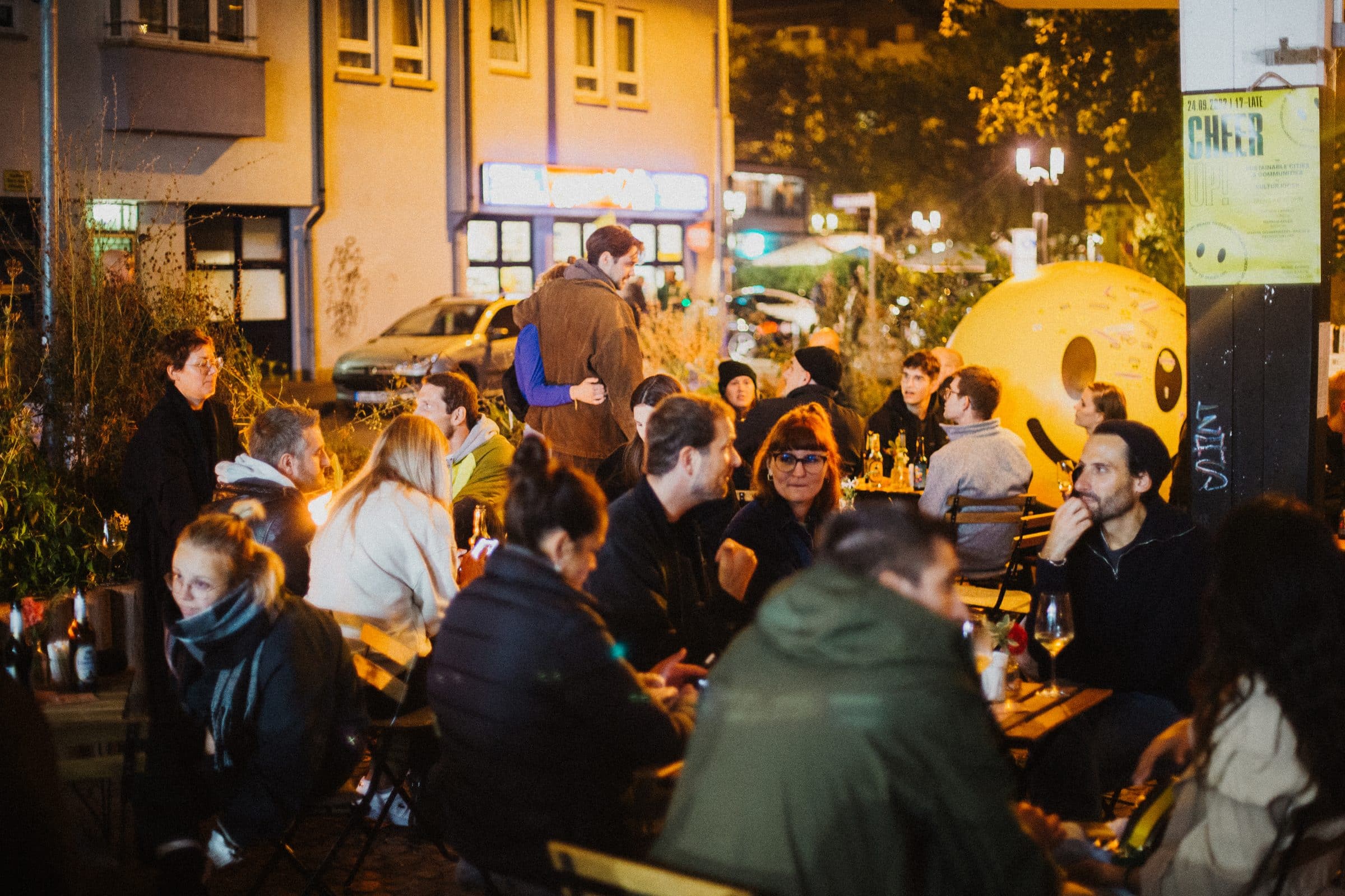 Guests sit at outdoor tables in the evening, with a yellow smiley-face tent in the background