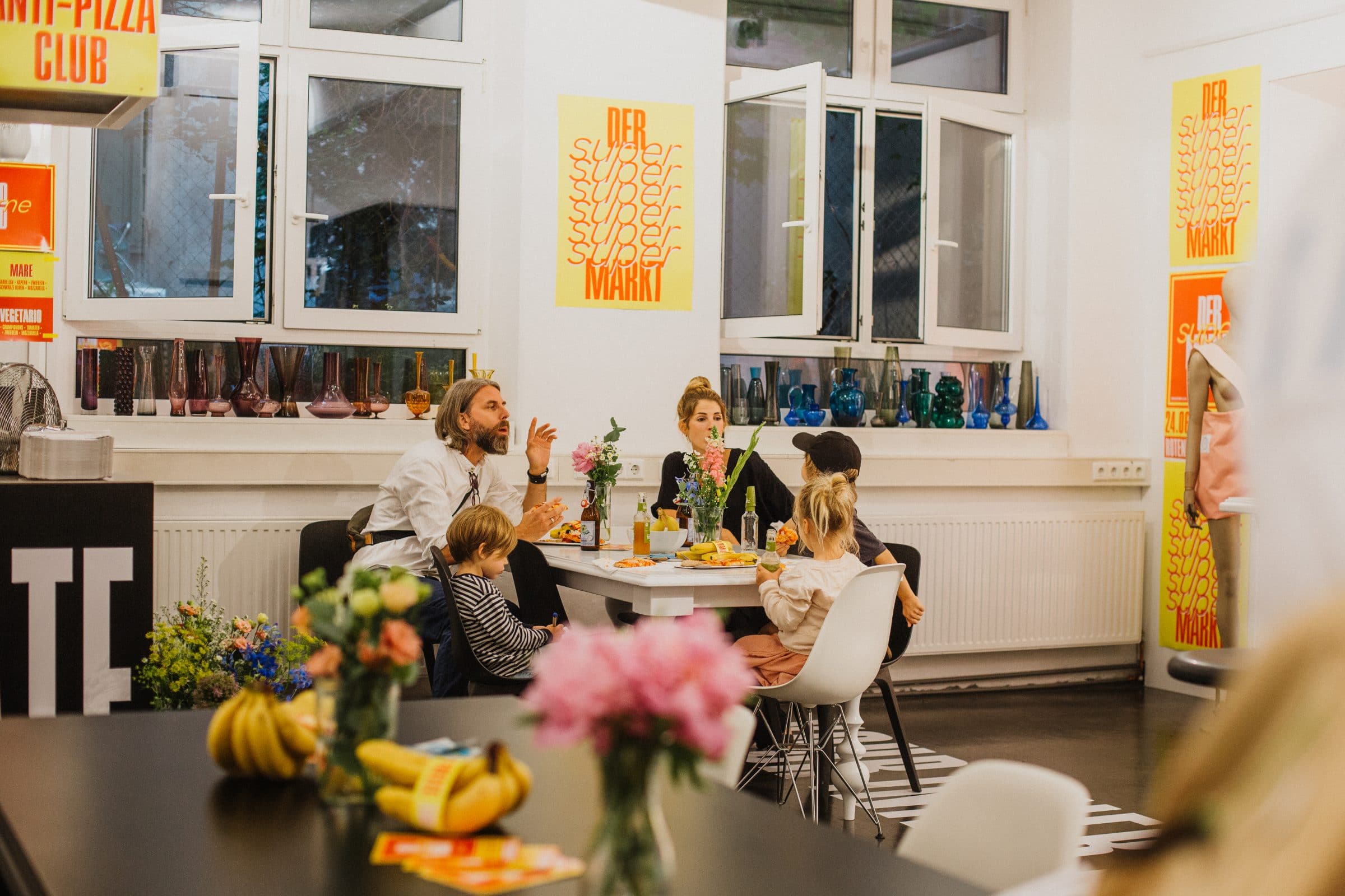 Familie sitzt am Tisch, im Hintergrund Poster „Der Super Markt“
