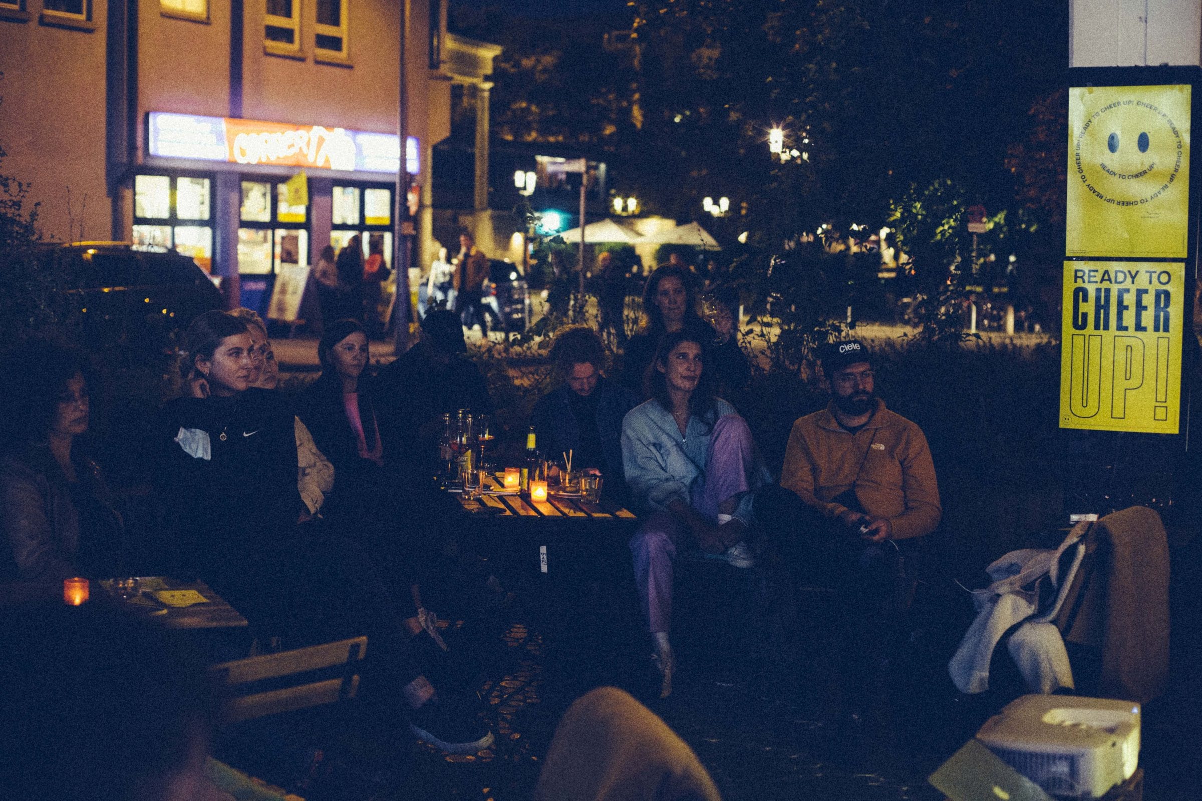 Audience seated outdoors at night in front of Kultur Kiosk, a yellow #CheerUpStuttgart poster behind them