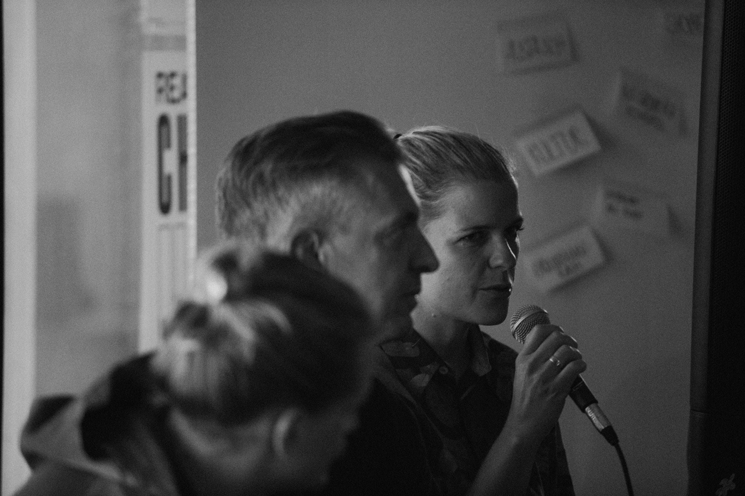 Black-and-white shot of a speaker with a microphone next to a moderator in front of a note-covered pinboard