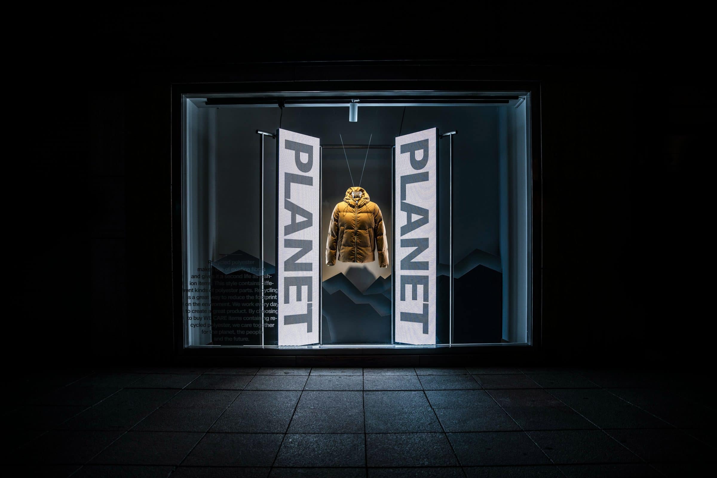 An illuminated nighttime shop window stages a yellow jacket between banners reading “PLANET”.