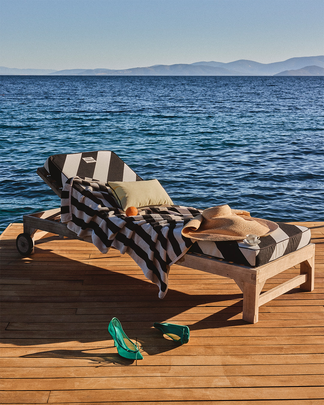 Jacquemus sun lounger with a striped towel, straw hat and green heels on a wooden deck by the sea