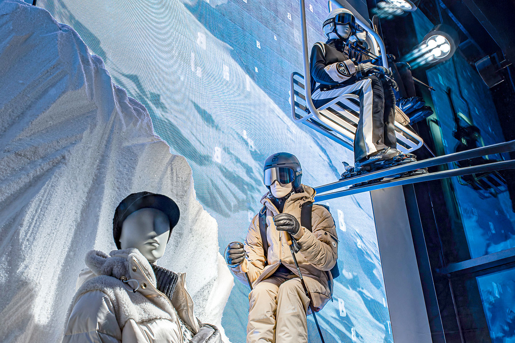 Nahaufnahme im BOGNER-Schaufenster: Mannequins in Skibekleidung vor Berglandschaft, darüber eine schwebende Gondel.