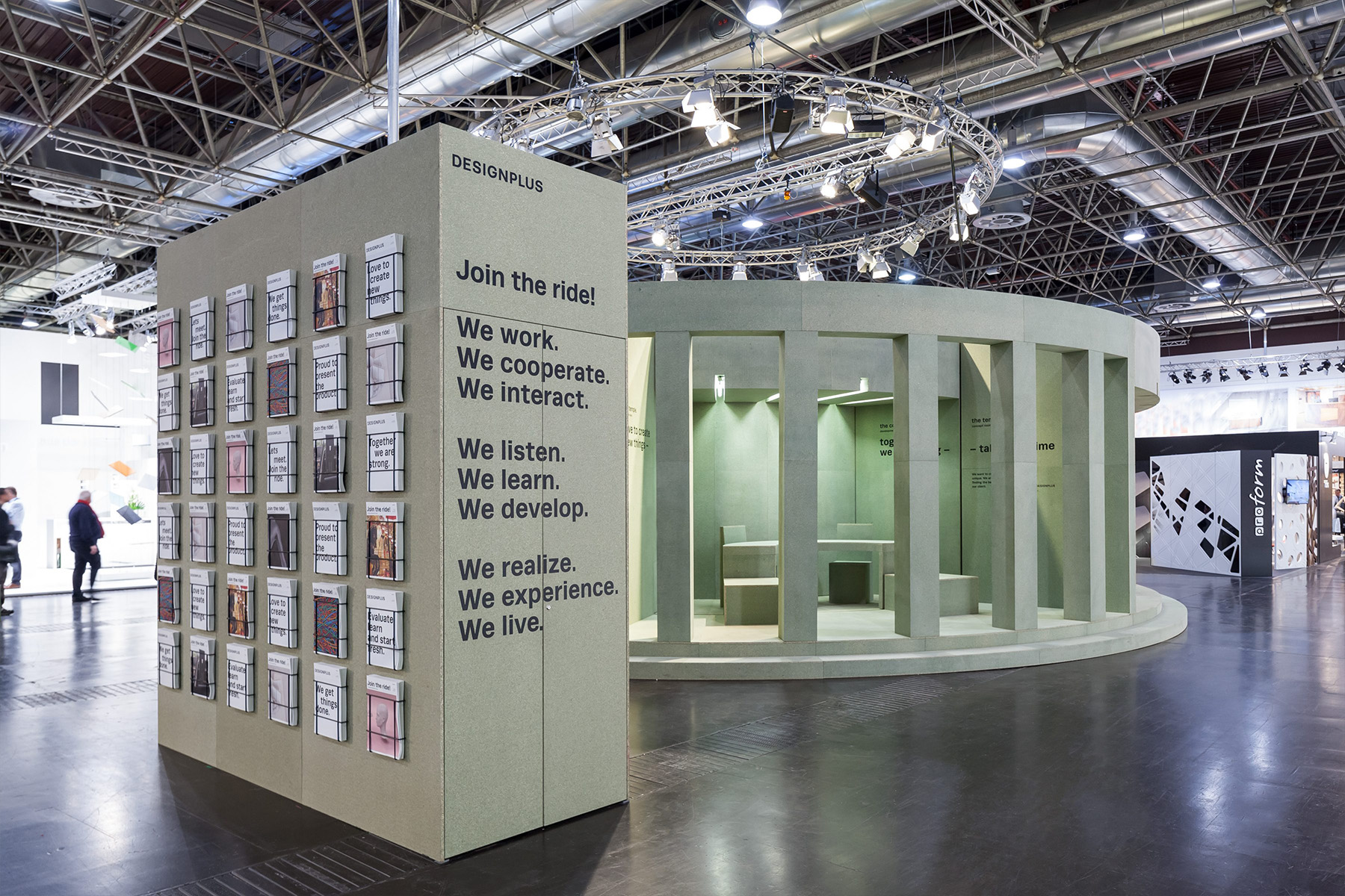 Der runde, drehbare Designplus-Messestand auf der EuroShop 2017 mit Journal-Wand und Sitznische, umgeben von Messehalle.