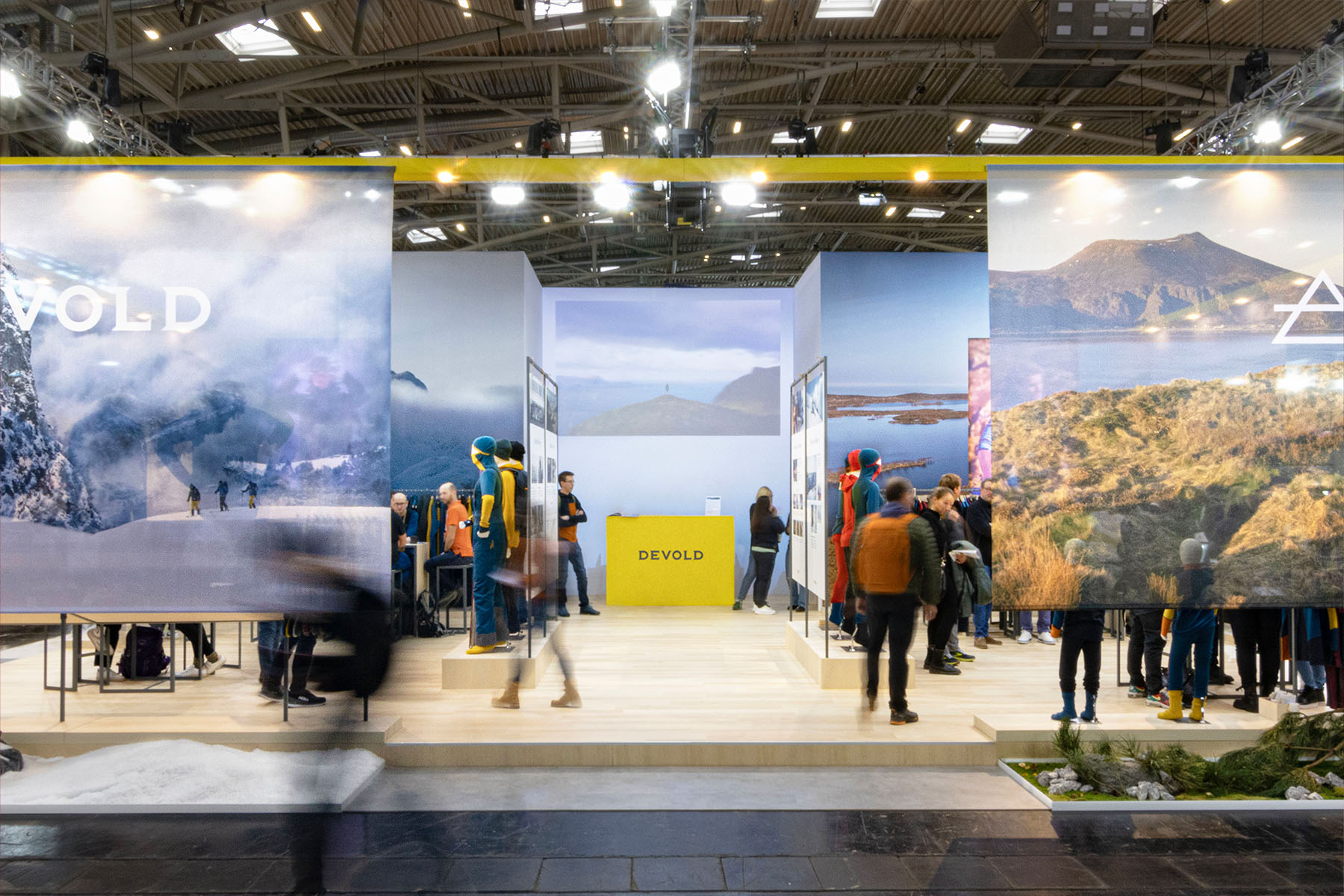 Devold-Messestand mit gelbem Empfangstresen, Landschaftsbannern und Besuchern auf Holzpodesten.