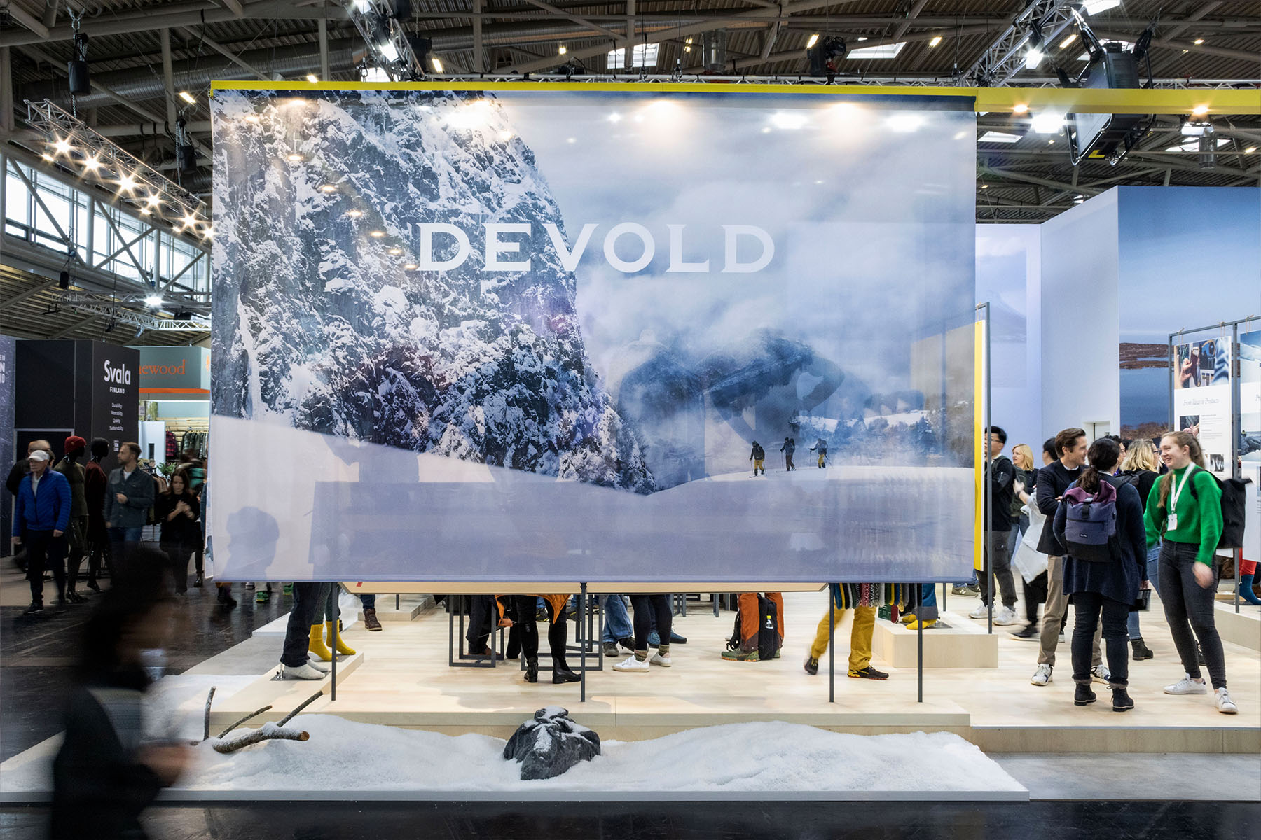Devold-Messestand: großformatiges Bergfoto-Banner mit Logo, darunter Besucher und Kunstschnee.