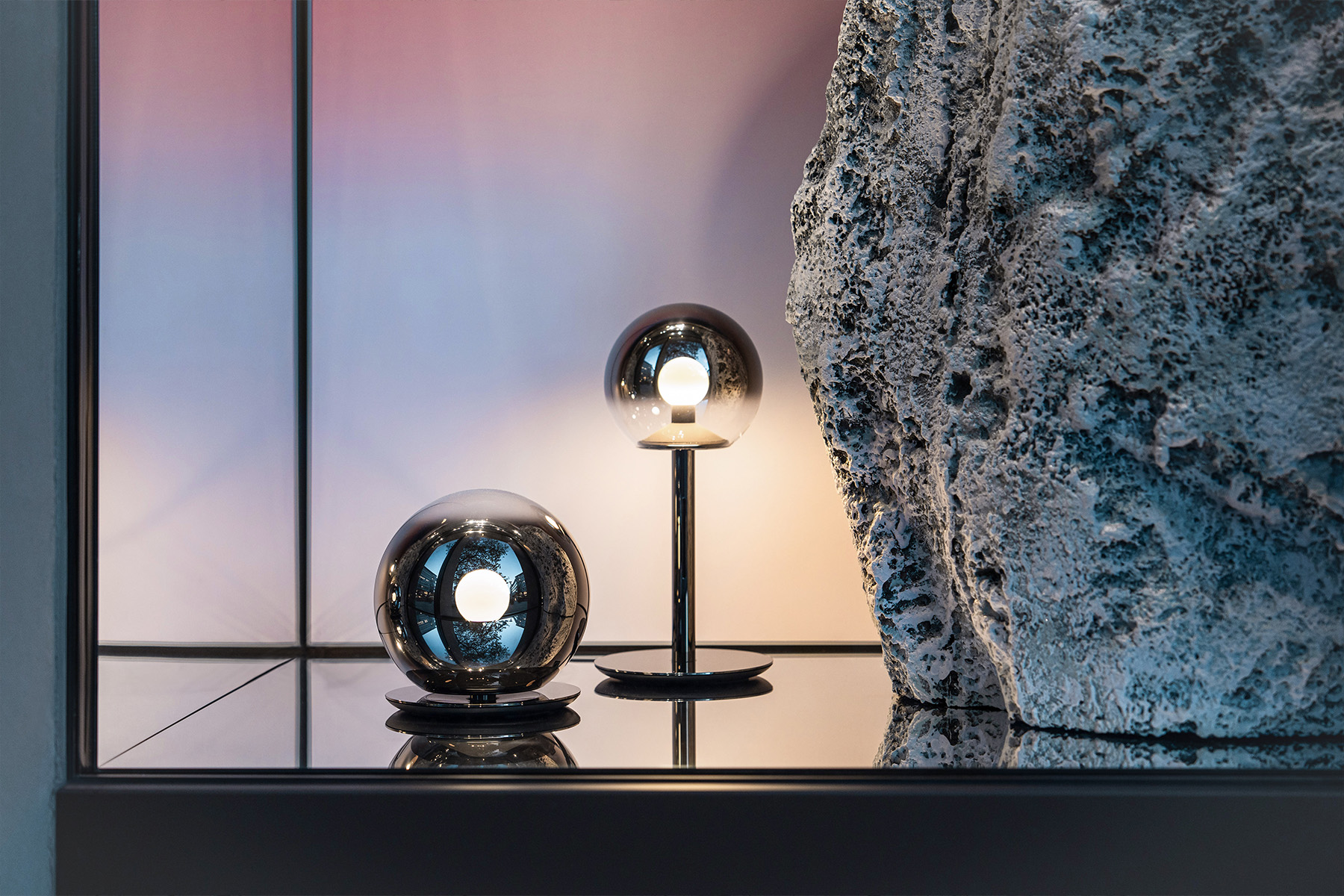 Two spherical Occhio table lamps glow next to a textured rock against a pastel gradient