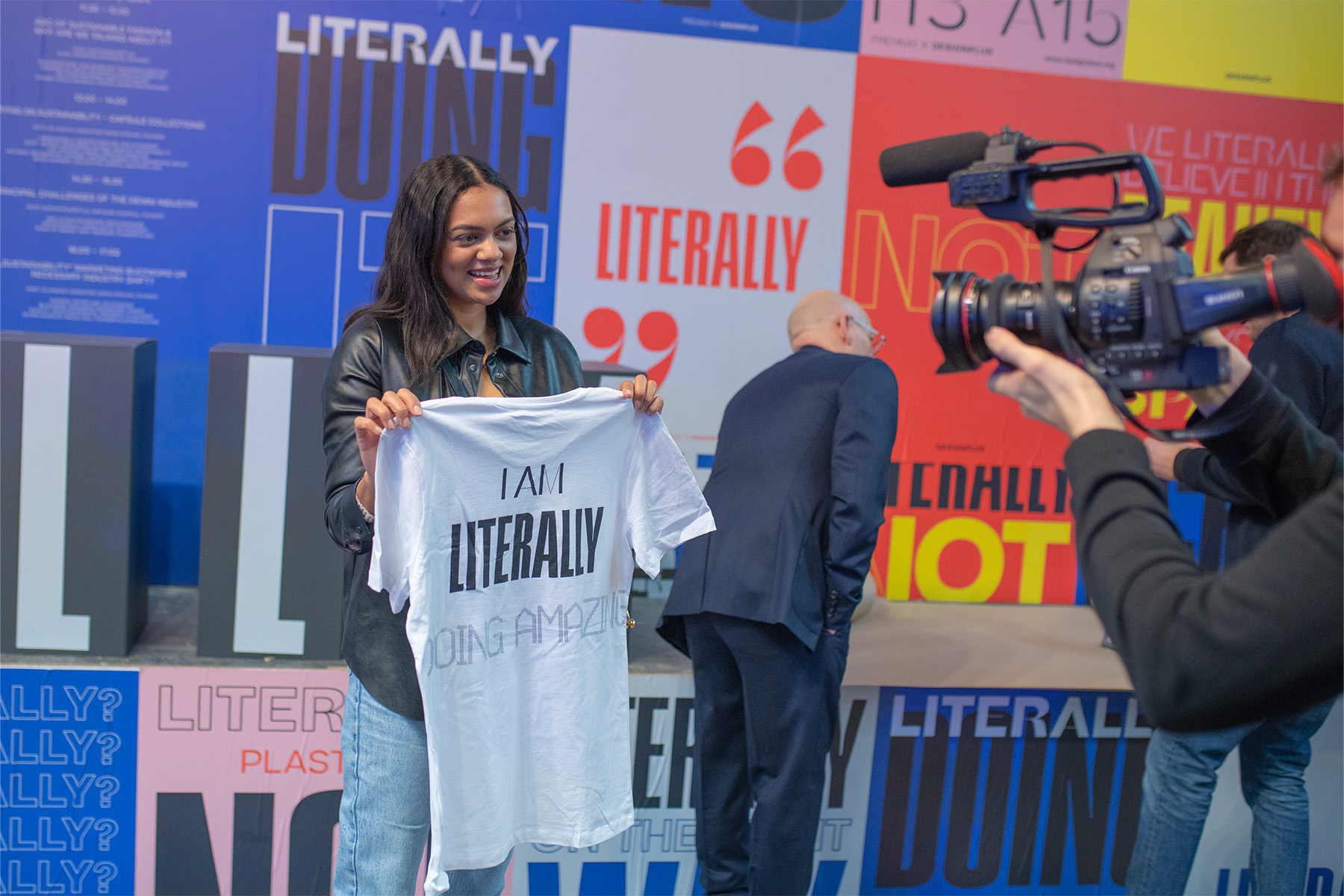 Frau präsentiert auf der Bühne ein T-Shirt mit „I Am Literally Doing Amazing“-Print.
