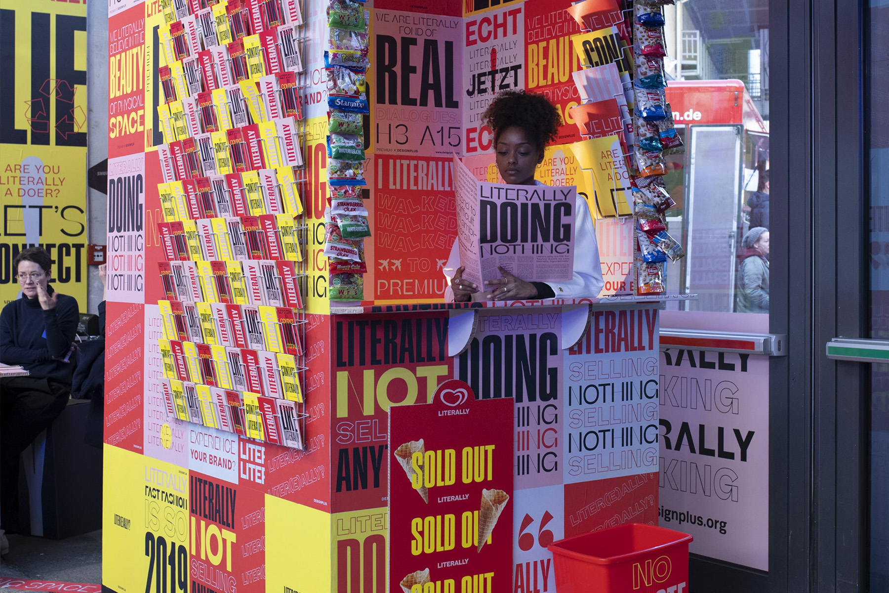Besucherin liest in einem bunten „Literally“-Kiosk mit Süßwaren- und „Sold Out“-Displays.