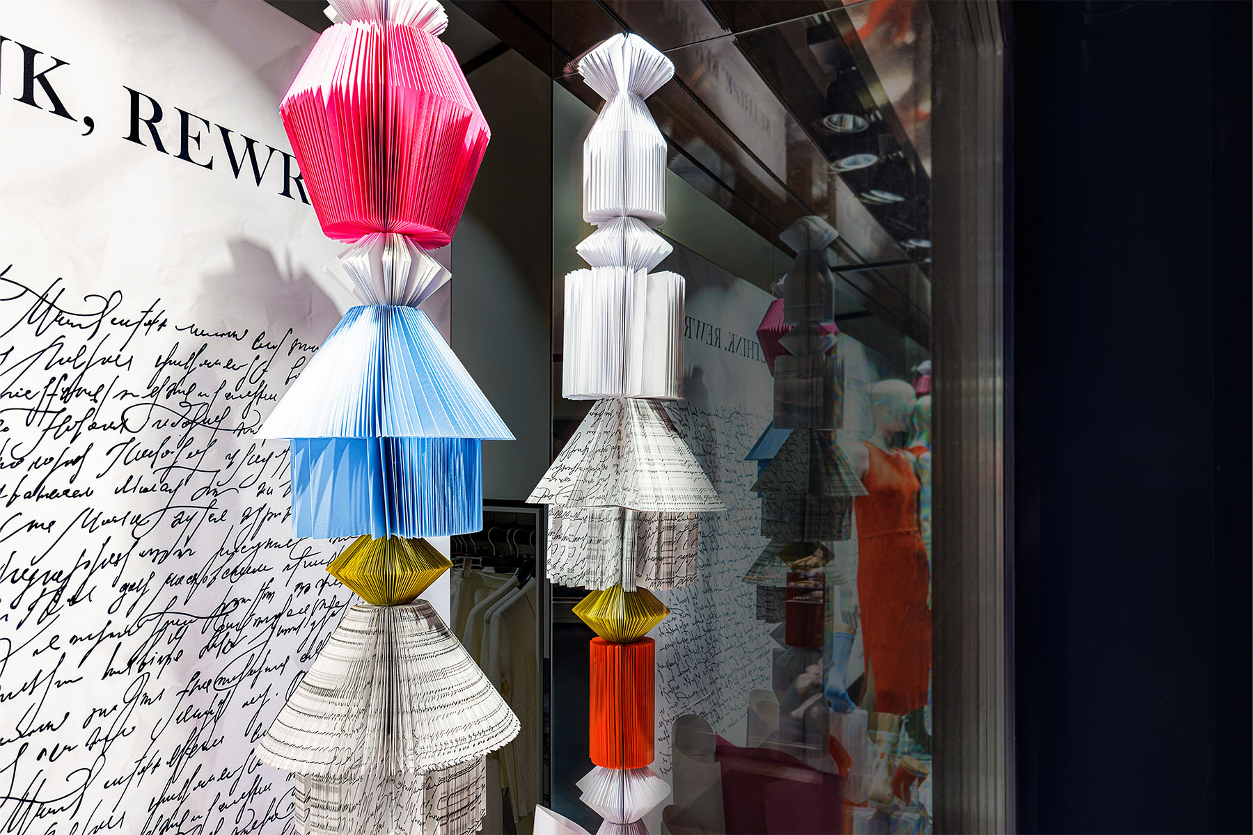 Colourful pleated paper sculptures in pink, blue and gold against handwritten text in the FALKE window