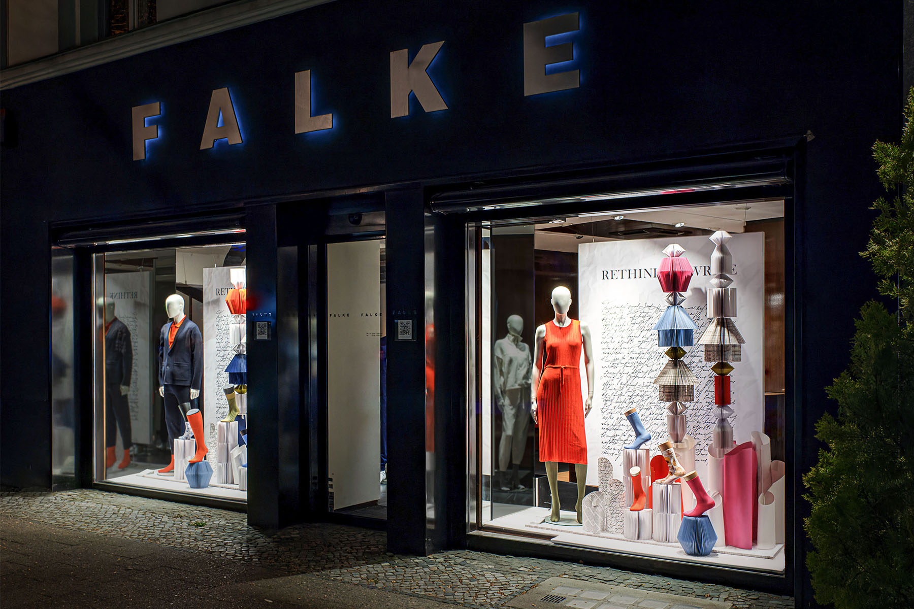 FALKE storefront at night with illuminated sign and two windows filled with paper sculptures