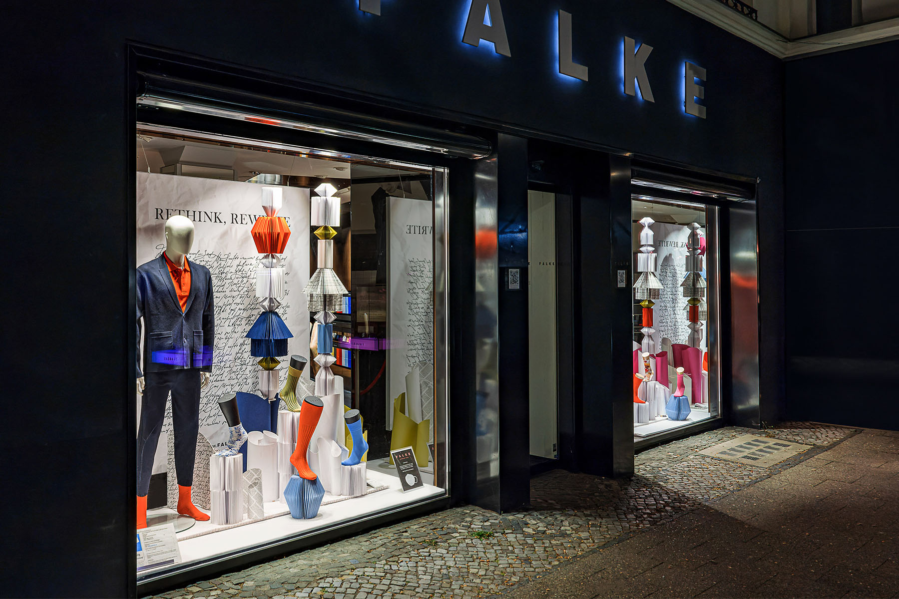 Angled night view of the FALKE storefront with illuminated displays and paper sculptures
