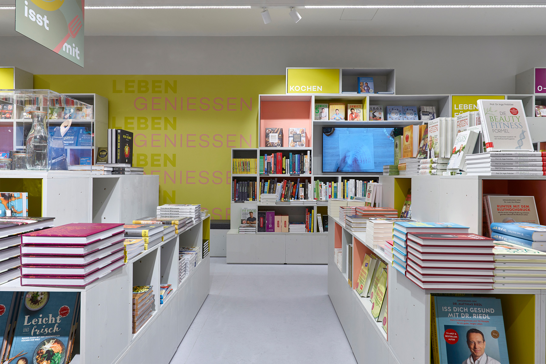 Hugendubel store interior with book tables, yellow wall typography reading "Leben Genießen" and the "Kochen" category sign.