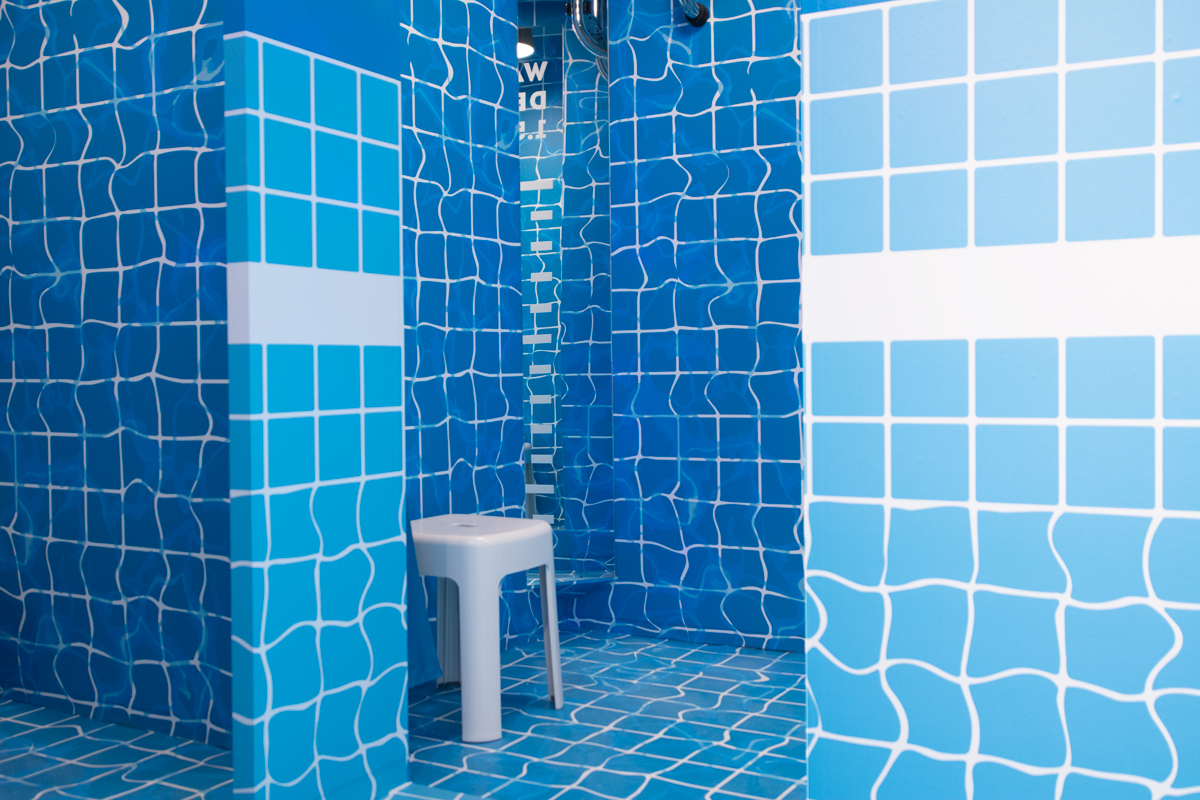 Umkleidekabine mit blauem Pool-Kachelmuster und weißem Hocker bei SportScheck.