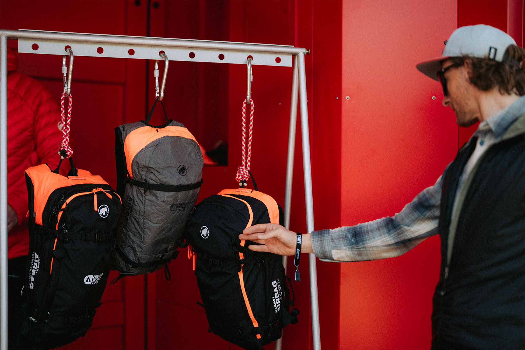 Besucher greift nach Mammut-Lawinenrucksäcken, die an einem Ständer vor rotem Container hängen.