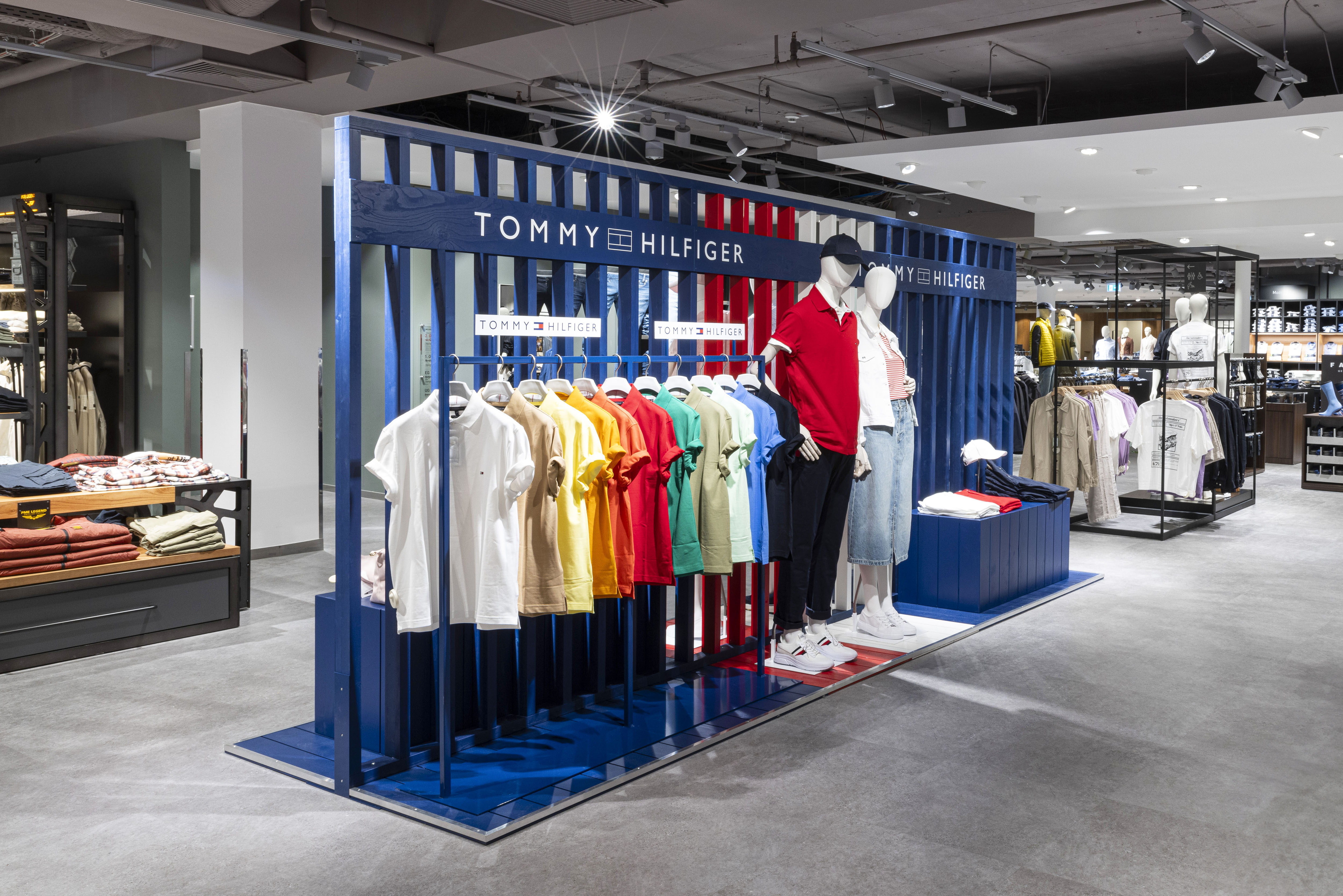 Tommy-Hilfiger-Shop-in-Shop mit blauem Holzgestell und Poloshirts in Regenbogenfarben.