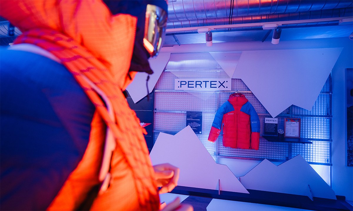 Rote Daunenjacke vor Pertex-Wand mit Berg-Grafiken, im Vordergrund Mannequin in Orange