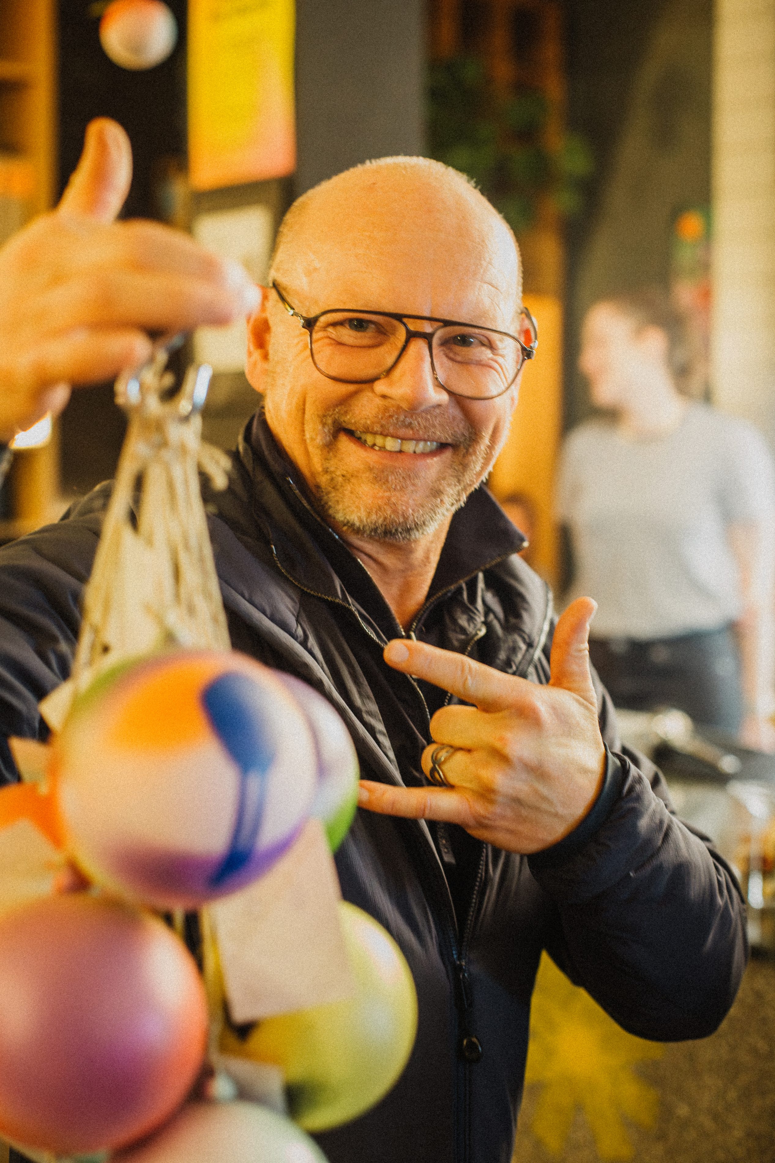 Mann hält lachend bunte Weihnachtskugeln in die Kamera und zeigt ein Peace-Zeichen