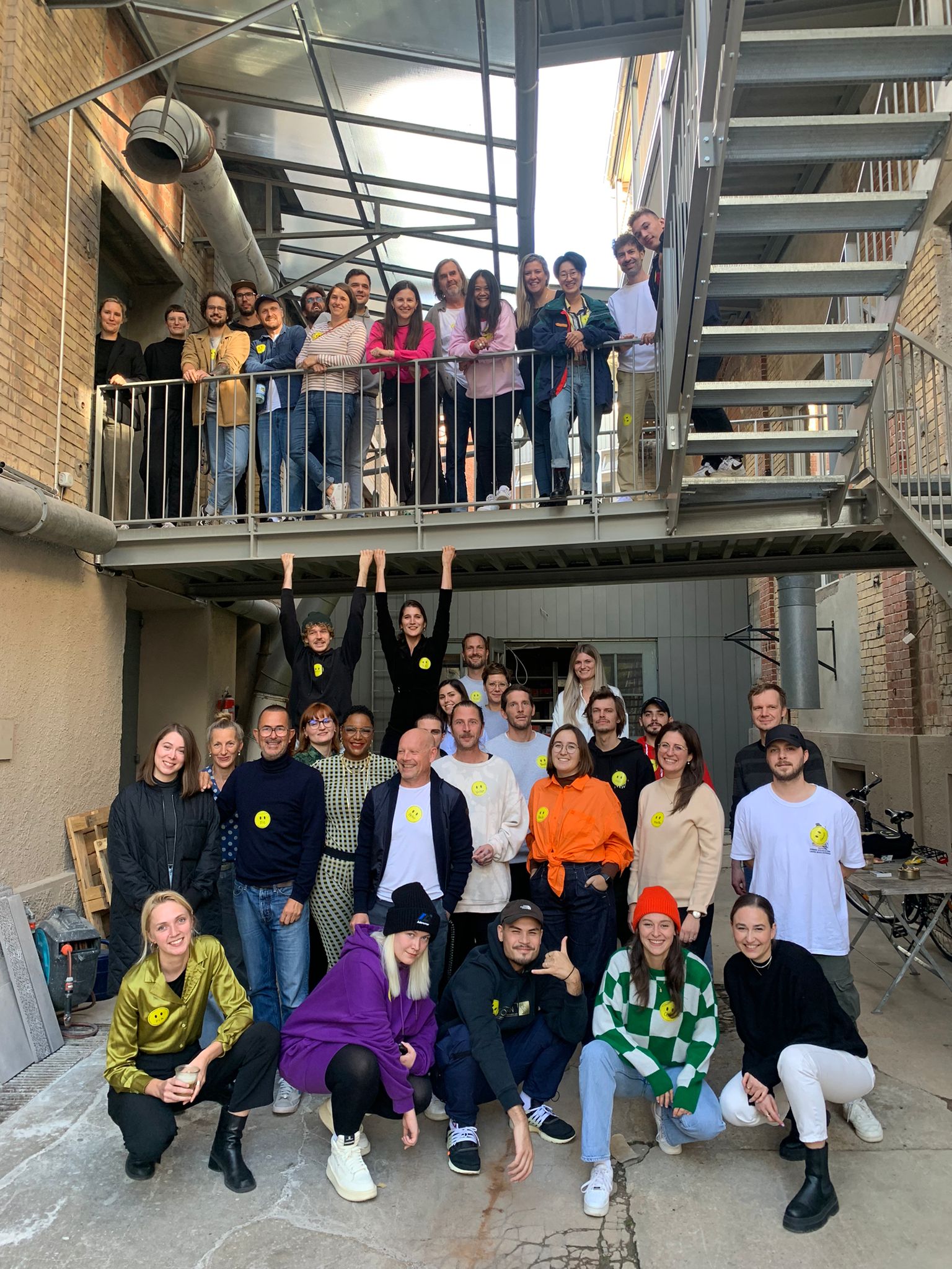 Gruppenfoto des gesamten Designplus-Teams auf der Außentreppe, alle mit gelben Smiley-Stickern