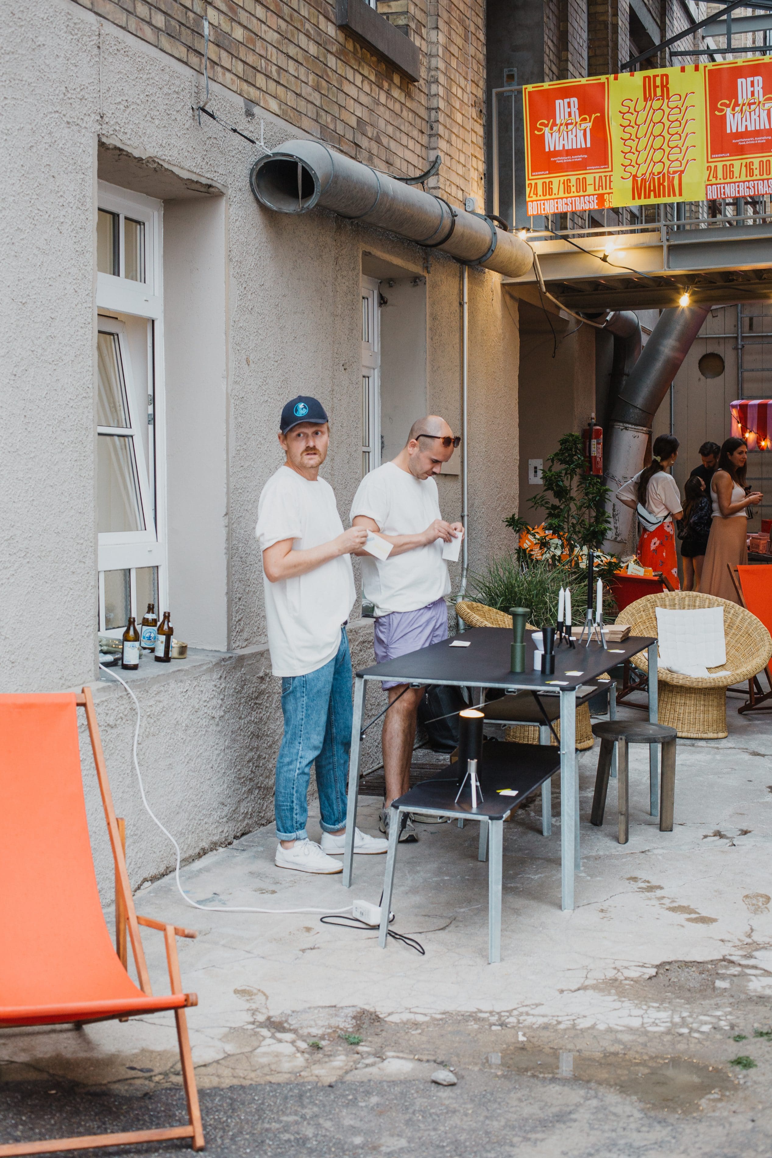 Two guests stand by a table, with “Der Super Markt” posters hanging above