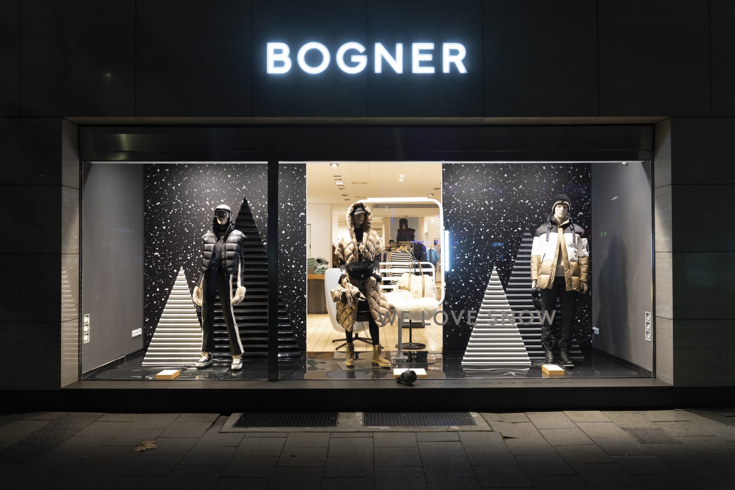 Bogner shop window at night: mannequins in ski wear in front of a snow motif with the “We love snow” lettering