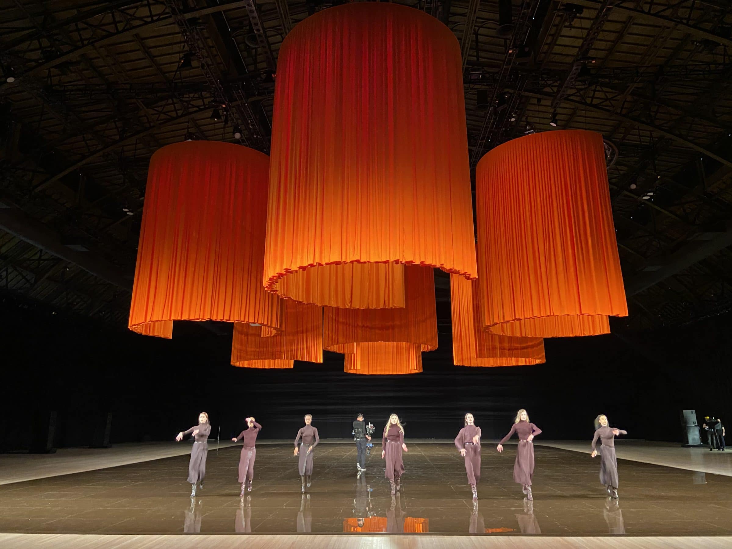 Giant orange fabric cylinders hang above the stage while performers dance below