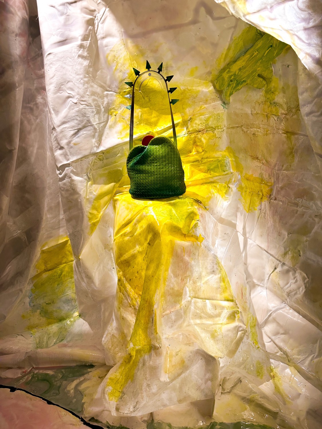 Green woven bag on a sculptural stand against a crumpled yellow-and-white backdrop