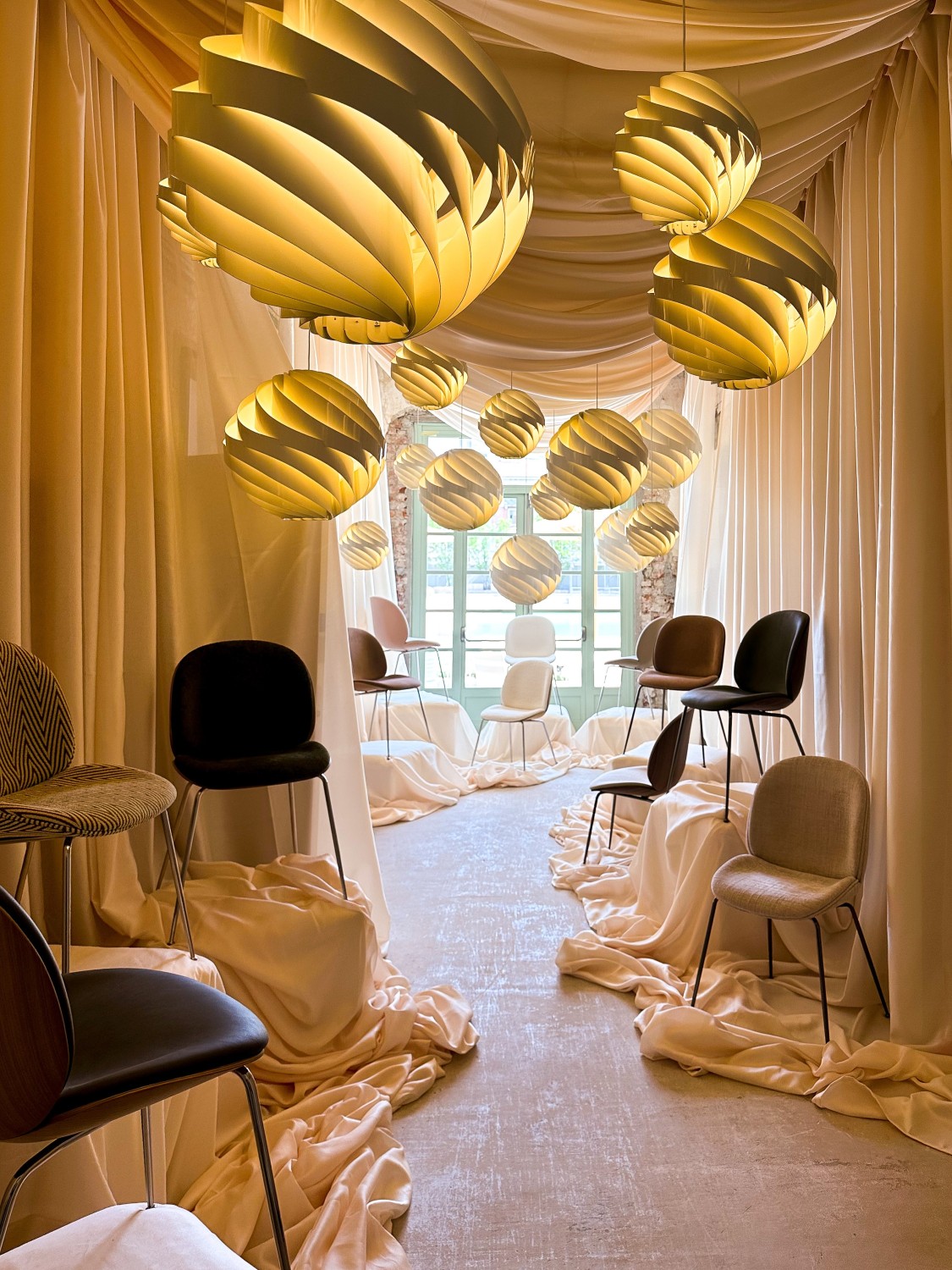 Exhibition room with golden globe pendant lights and Beetle chair variants on draped fabric, GUBI installation