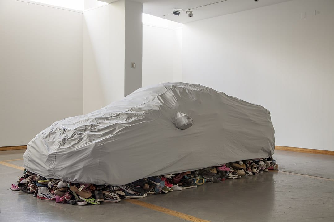 Skulptur „Fortified“ von Karyn Olivier: autoförmige graue Hülle auf einem Bett aus alten Schuhen