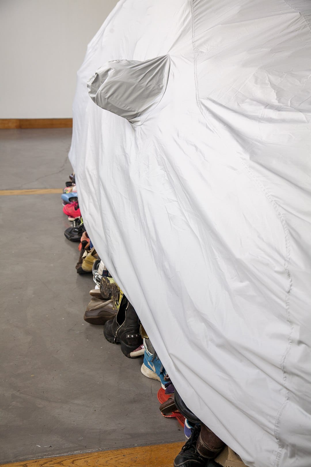 Skulptur von Karyn Olivier: grauer Autoüberzug über einem Objekt, darunter zahlreiche Schuhe hervorschauend