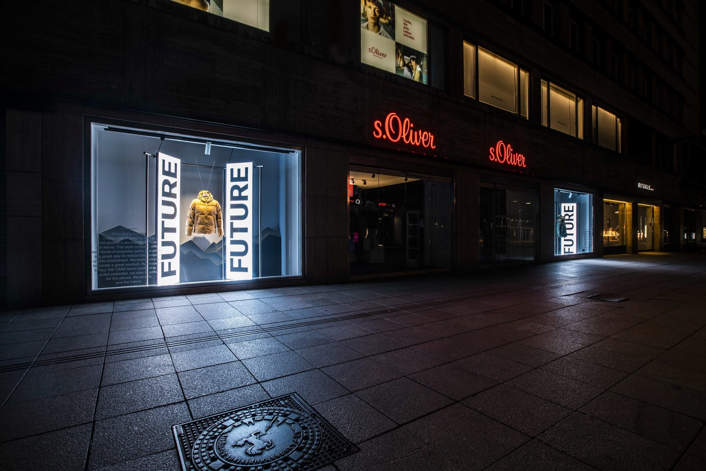 Beleuchtetes s.Oliver-Schaufenster bei Nacht mit Jacke zwischen „FUTURE“-Bannern, im Hintergrund die rote Filialbeschriftung.