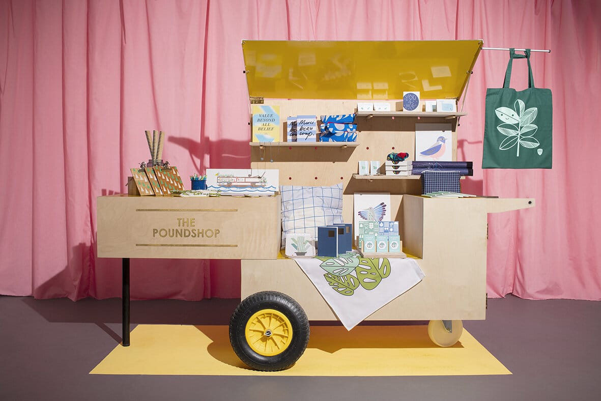 Mobiler Verkaufswagen „The Poundshop“ aus Holz mit gelbem Rad vor rosa Vorhang