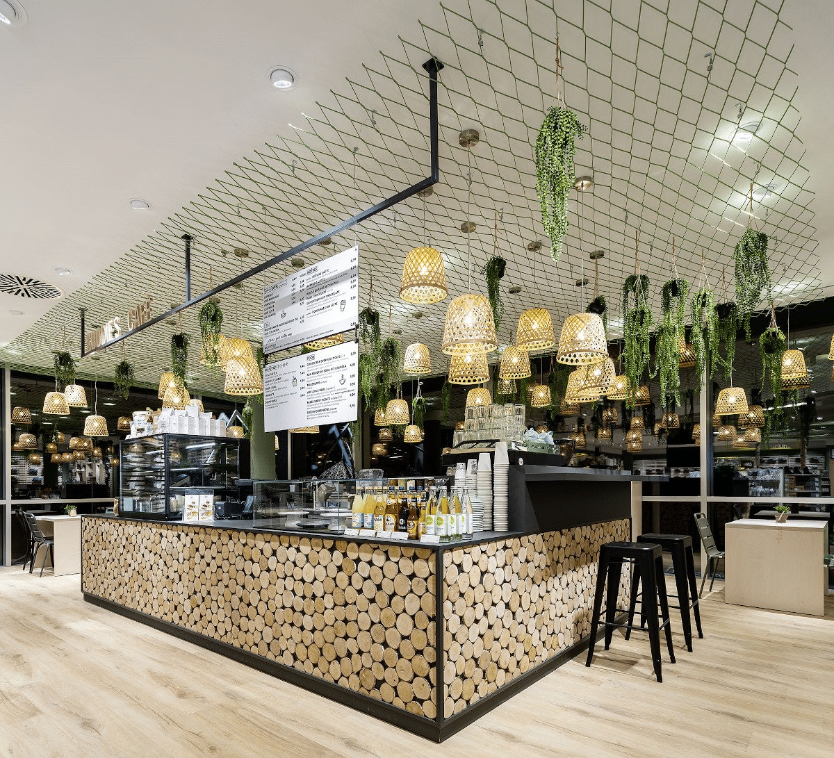 A café counter clad in log-slice tiles beneath hanging rattan lampshades and greenery.