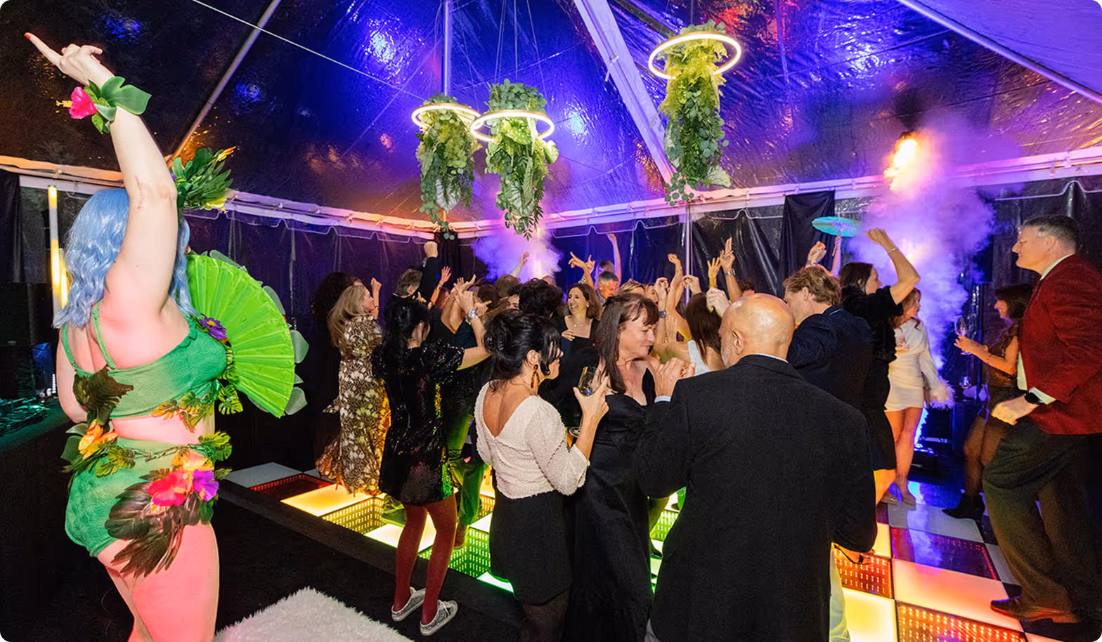 Packed dance floor under a tent with purple lighting, hanging greenery and a costumed performer