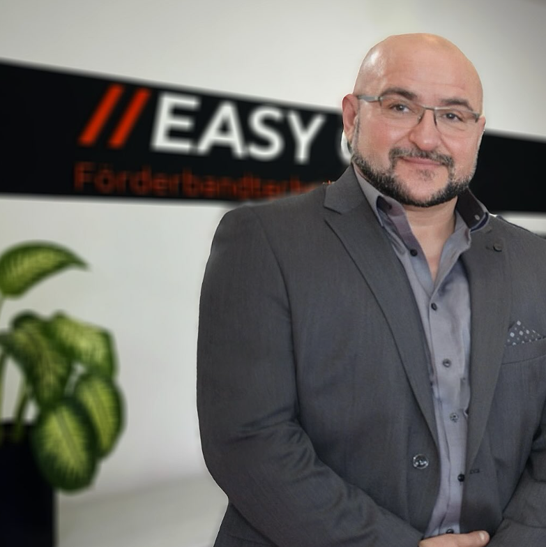 Smiling bald man with glasses and a beard wearing a gray suit and shirt standing indoors.