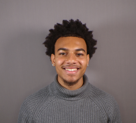Smiling man with short curly hair wearing a gray knitted turtleneck sweater in front of a plain gray background.